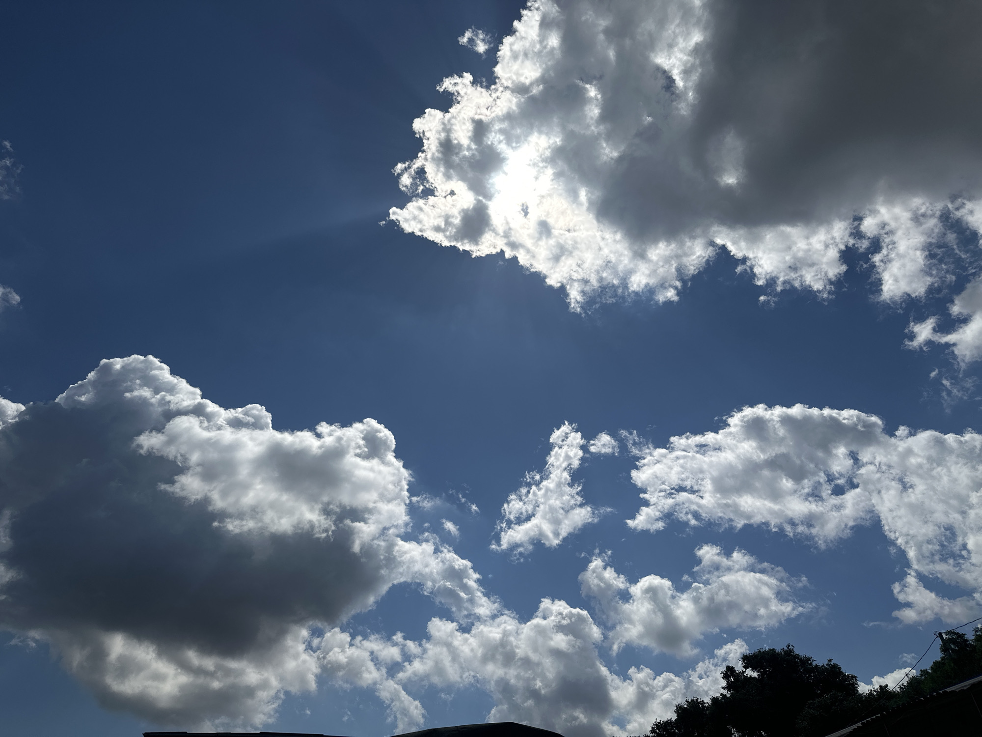 The image depicts a sky filled with dramatic clouds and sunlight. In the upper portion of the frame, the sun is partially obscured by a large, bright cloud, casting rays of light downwards. Below this, a darker, swirling cloud formation is visible. The lower half of the image shows more scattered cumulus clouds against a clear blue sky. Silhouetted against the sky at the very bottom are the tops of trees and what appears to be the edge of a roof. The overall scene is natural and depicts a dynamic sky. There are no people or specific identifiable landmarks that would suggest a location in the city of Taman, Russia.