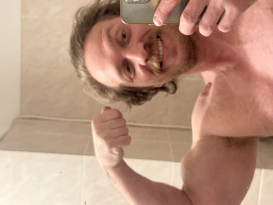 A man, topless, flexing his right bicep and smiling at his reflection in a bathroom mirror. He's holding a phone to take the picture. He has curly hair and a mustache. The bathroom has off-white walls and a molding or shelf visible along the bottom of the mirror. The image is taken at an unusual angle, with the man looking down into the mirror.