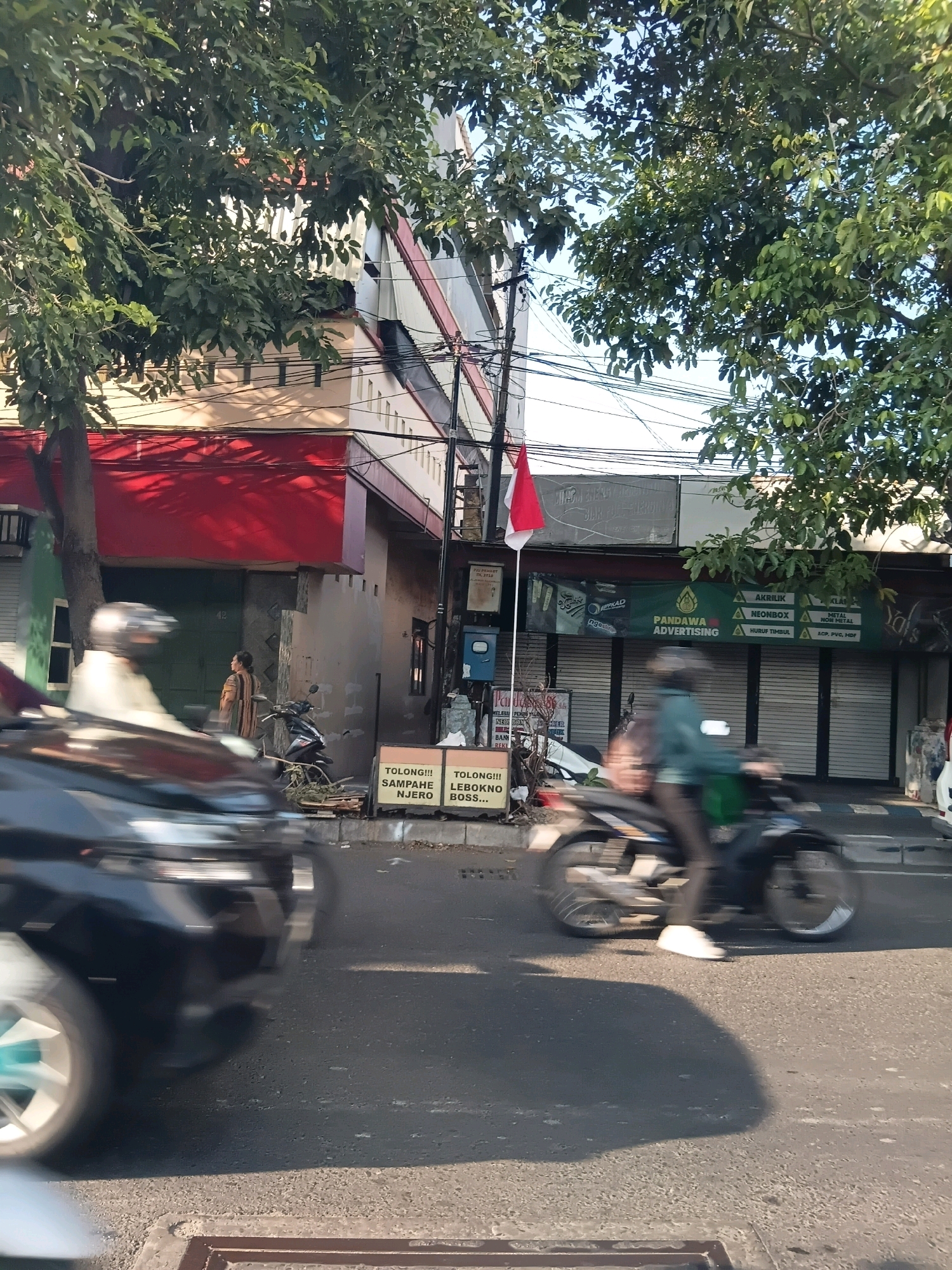 The image captures a street scene in Surabaya, Indonesia, with buildings lining one side of the road. A large tree with dense green foliage partially obscures the upper portion of the buildings. In the foreground, a black car is blurred due to motion, indicating it is in transit. To the right, a person on a motorcycle, also blurred by motion, is riding down the street. The motorcycle is dark colored. A few other people are visible further down the street, one standing near a building entrance and another person on a scooter, partially visible. There are several signs on the buildings, including one with "PANDAWA ADVERTISING" written on it. A red and white Indonesian flag is flying from a pole. The road surface appears to be asphalt, and a drain grate is visible in the lower foreground. The lighting suggests it is daytime, with sunlight casting shadows.