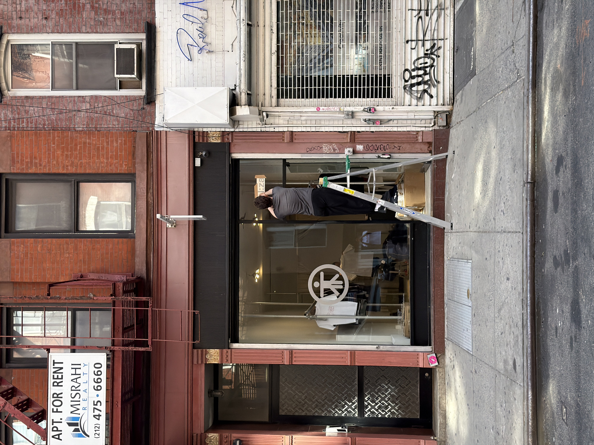 The image shows the facade of a building in New York City, United States. The building has a storefront with a large glass window. A man is inside the storefront, standing on a ladder and appearing to be working on something near the top of the window. Through the glass, clothing can be seen in the retail space. The storefront window has a logo on it. Above the storefront, there is a security gate covering a window. The building is brick and has a fire escape on the left side. A sign on the fire escape advertises apartments for rent with the phone number (212) 475-6660. The building is adjacent to a sidewalk.
