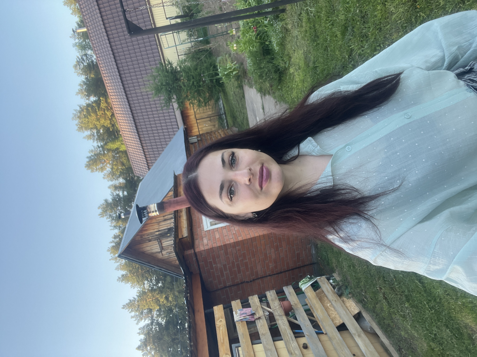 The image shows a woman with long, dark hair wearing a light blue, polka-dotted blouse. She is outdoors, with a house visible in the background. The house appears to be a brick structure with a dark roof and a chimney. The environment appears to be a yard or garden with greenery and a wooden fence. The image was likely taken in Garevaya, Russia.