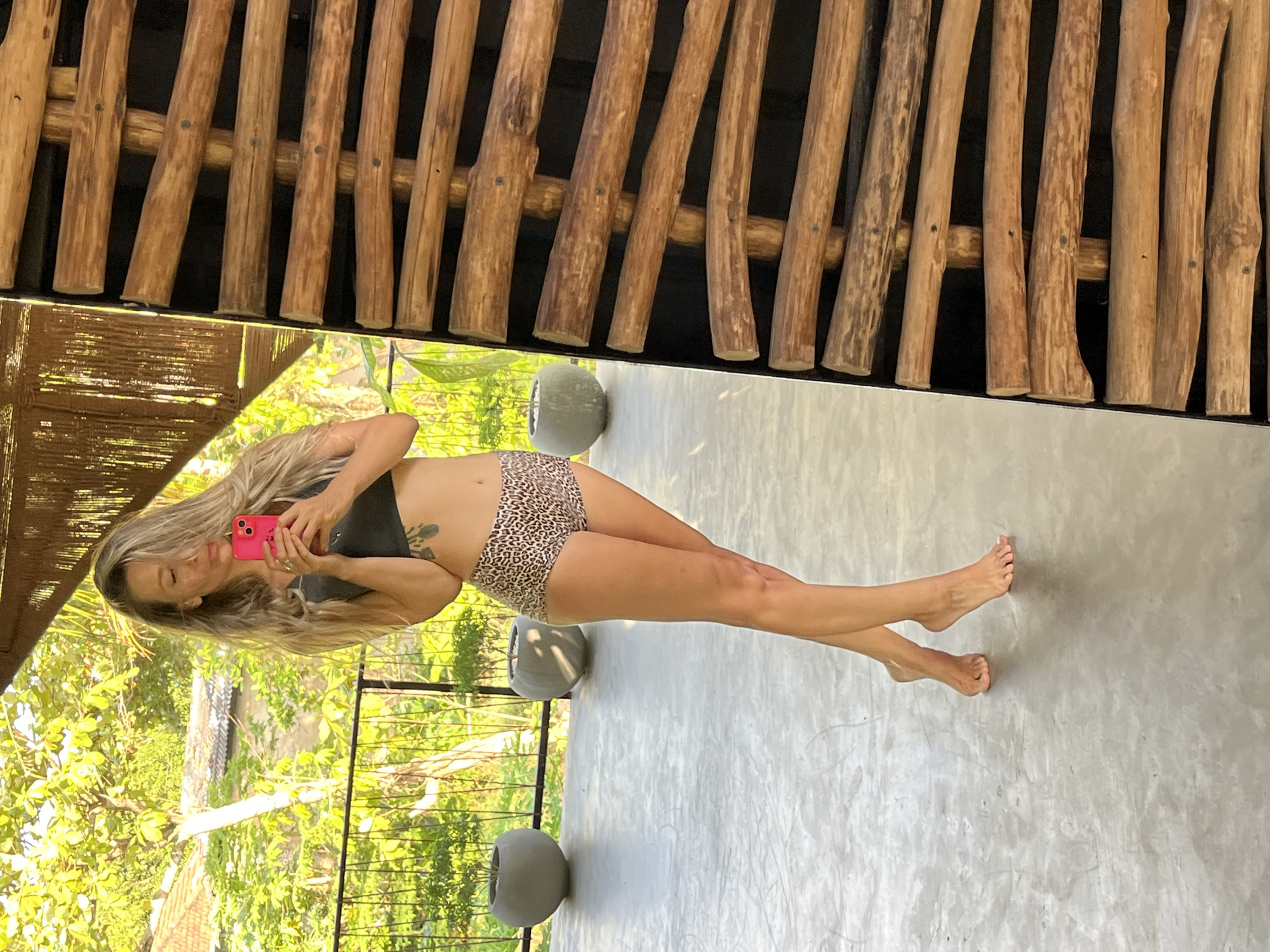 A woman stands on a concrete floor, facing a mirror, taking a selfie with her pink phone. She is wearing a dark grey crop top and leopard print shorts. Her long blonde hair is down and appears windswept. Behind her, through a railing and lush green foliage, are glimpses of buildings with terracotta roofs. Two large grey planters are visible in the background. The right side of the image is dominated by a textured wall made of horizontally stacked wooden logs. The scene suggests an outdoor or semi-outdoor setting, possibly a balcony or patio area, with a tropical ambiance. The location is identified as Weligama, Sri Lanka.