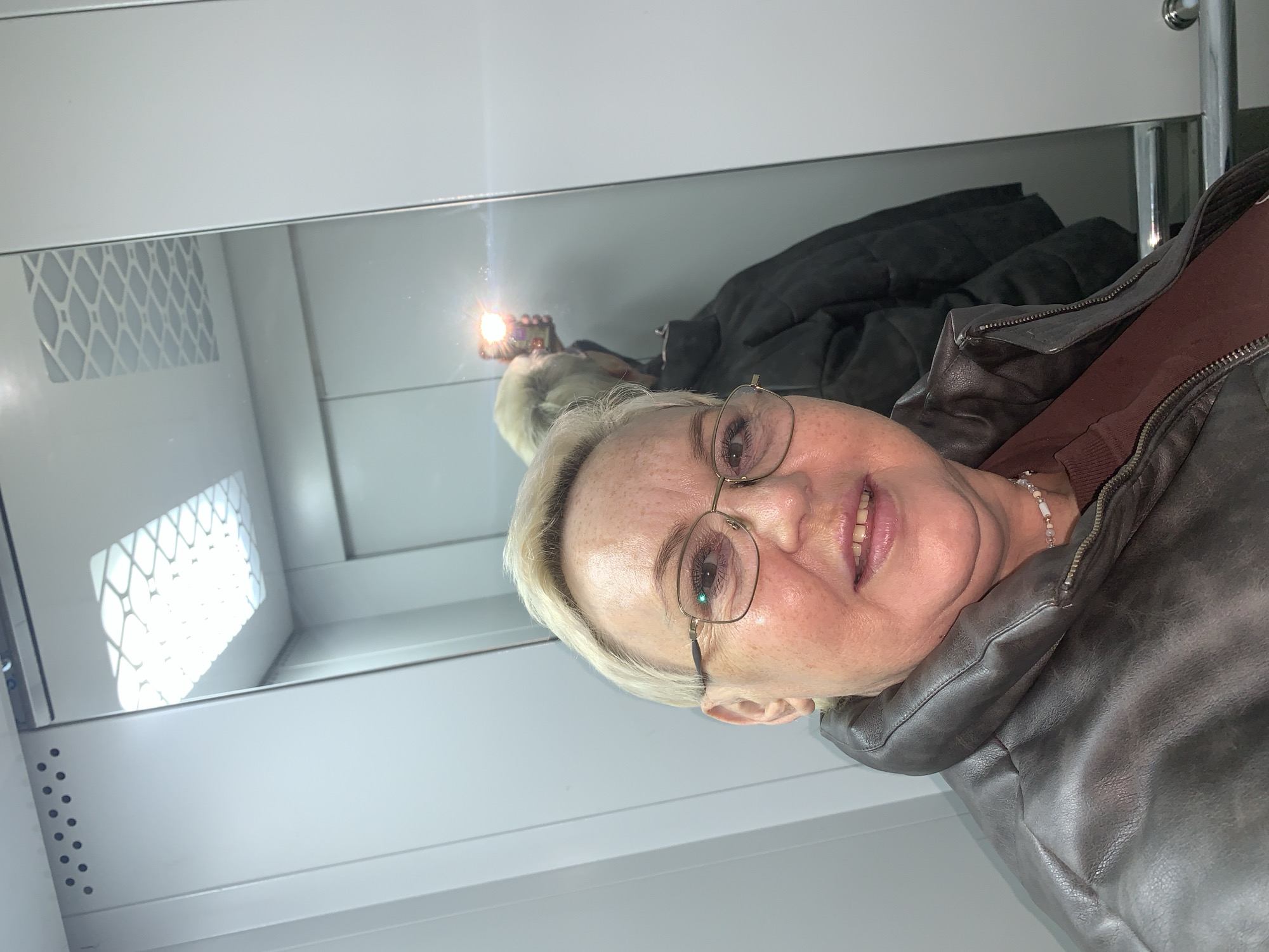 An image taken in an elevator in the city of Chaikovsky, Russia, features a middle-aged woman taking a selfie. The woman, with short, light blonde hair and a faint smile, occupies the left half of the frame, facing the camera directly. She wears gold-rimmed rectangular eyeglasses, which show a green reflection in the right lens. Her attire includes a dark brown leather-like jacket with a prominent collar, unzipped to reveal a reddish-brown garment underneath. A light-colored beaded necklace is visible around her neck.
To the right, a large rectangular mirror reflects the elevator's interior and the woman's back. In this reflection, her short blonde hair and the back of a dark, possibly padded jacket (distinct from the one she is wearing in the foreground) are visible. A bright, circular flash of light, emanating from what appears to be a smartphone held by her reflected arm, is prominently displayed in the mirror. The ceiling of the elevator, featuring a rectangular light fixture with a geometric grid pattern, is also reflected in the mirror. The elevator walls are light grey or white, with vertical paneling. On the upper left wall, a horizontal row of small, dark circular vents is present. A metallic horizontal bar, likely a handrail, is partially visible at the bottom right of the mirror's reflection.