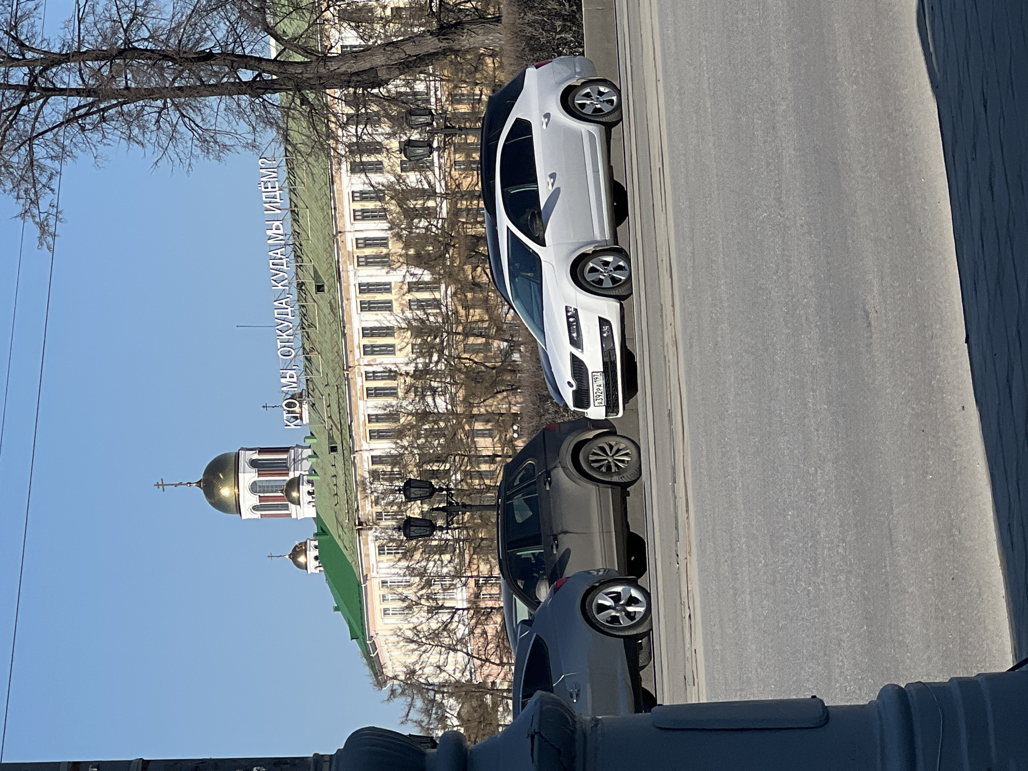 The image captures a sunny street scene in Yekaterinburg, Russia, likely during late winter or early spring, indicated by the bare tree branches. The perspective is slightly elevated, looking down upon a multi-lane road.
In the foreground, a dark railing or architectural element is visible on the right. On the grey asphalt road, three cars are present. Closest to the viewer is a dark-colored car, followed by a dark SUV, and then a white hatchback, possibly a Skoda Octavia, with a black roof. The white car bears a Russian license plate: "A392PA 197".
Behind the road, a large, ornate building with light-colored facades and numerous windows stretches across the midground. Its green roof is prominent. Rising above this building are two golden domes, topped with Orthodox crosses, signifying a church structure. Bare tree branches frame the upper left portion of the scene. Several classic black street lamps are positioned between the road and the main building.
A striking detail is the large, white Russian text displayed on the green roof of the main building, which reads: "КТО МЫ, ОТКУДА, КУДА МЫ ИДЁМ?" This translates to "Who are we, where from, where are we going?", posing a philosophical question as a public art installation or message. The sky is clear blue, indicating bright daylight. The scene depicts a typical urban environment blending historical architecture with modern street life and an intriguing intellectual element.