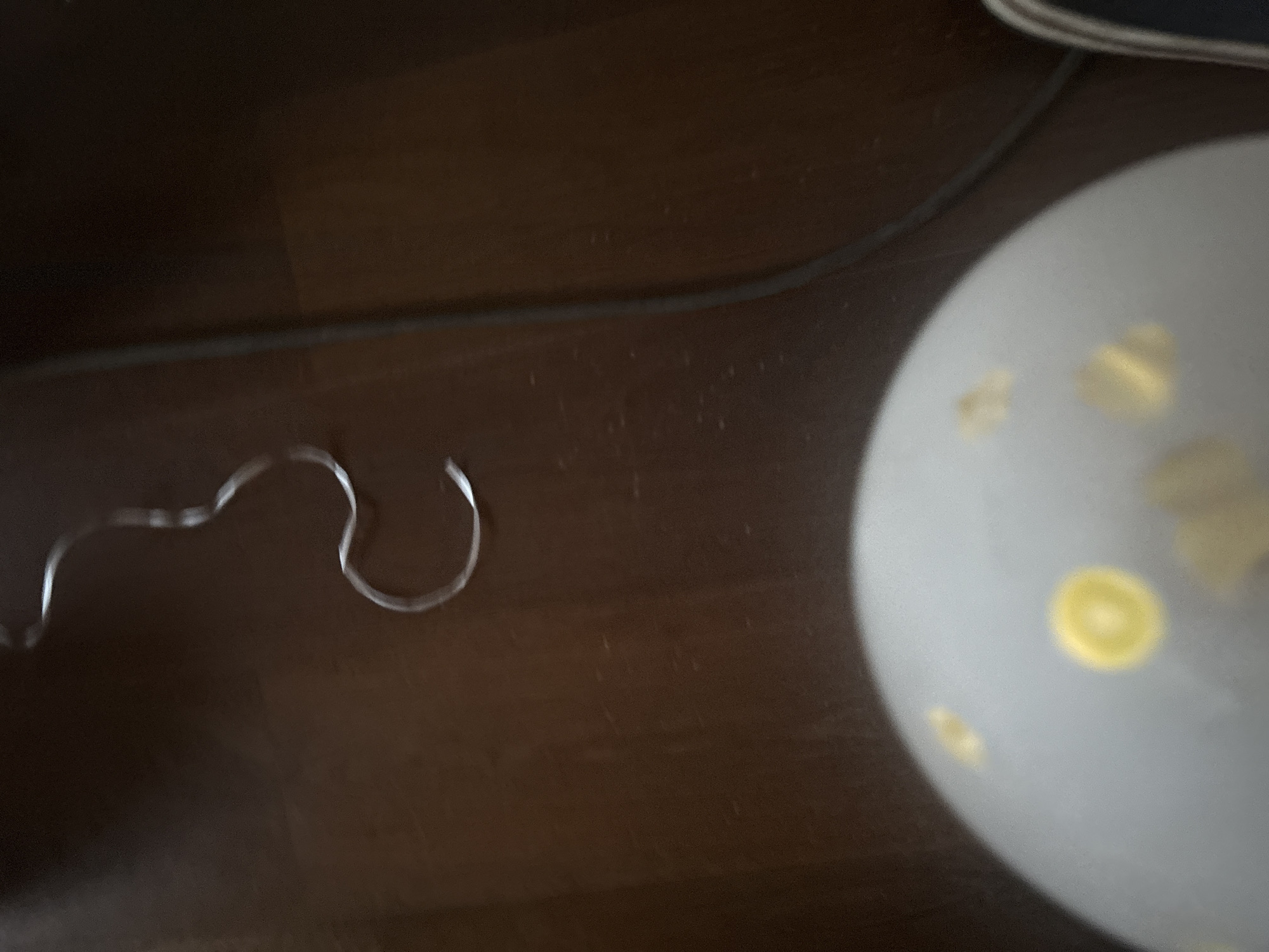 The image shows a close-up, high-angle view of a light gray spherical object, possibly a balloon, with yellow circular and irregular shapes on its surface. The object is positioned on a dark brown wooden floor. To the upper right of the balloon, a thin, silver ribbon is visible, bent into a curved shape. A dark gray cable runs diagonally from the top right to the bottom right of the frame, also resting on the wooden floor. The background is predominantly the dark brown floor. No people or interactions are depicted. The scene is static. This image does not provide any information about the city of Orekhovka, Russia, or any other geographical location.