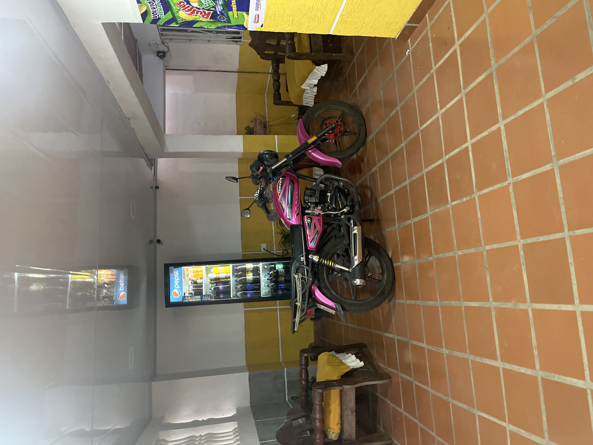 The image shows the interior of a commercial establishment, likely a small store or shop, in Juan Griego, Venezuela. The flooring is tiled in terracotta squares. The left side of the image shows a white-walled area with a highly reflective floor, likely polished tile. A refrigerated display case filled with beverages is visible against a light yellow wall. A pink motorcycle is parked centrally, its tires facing different directions. Wooden chairs and a small wooden bench are also present.