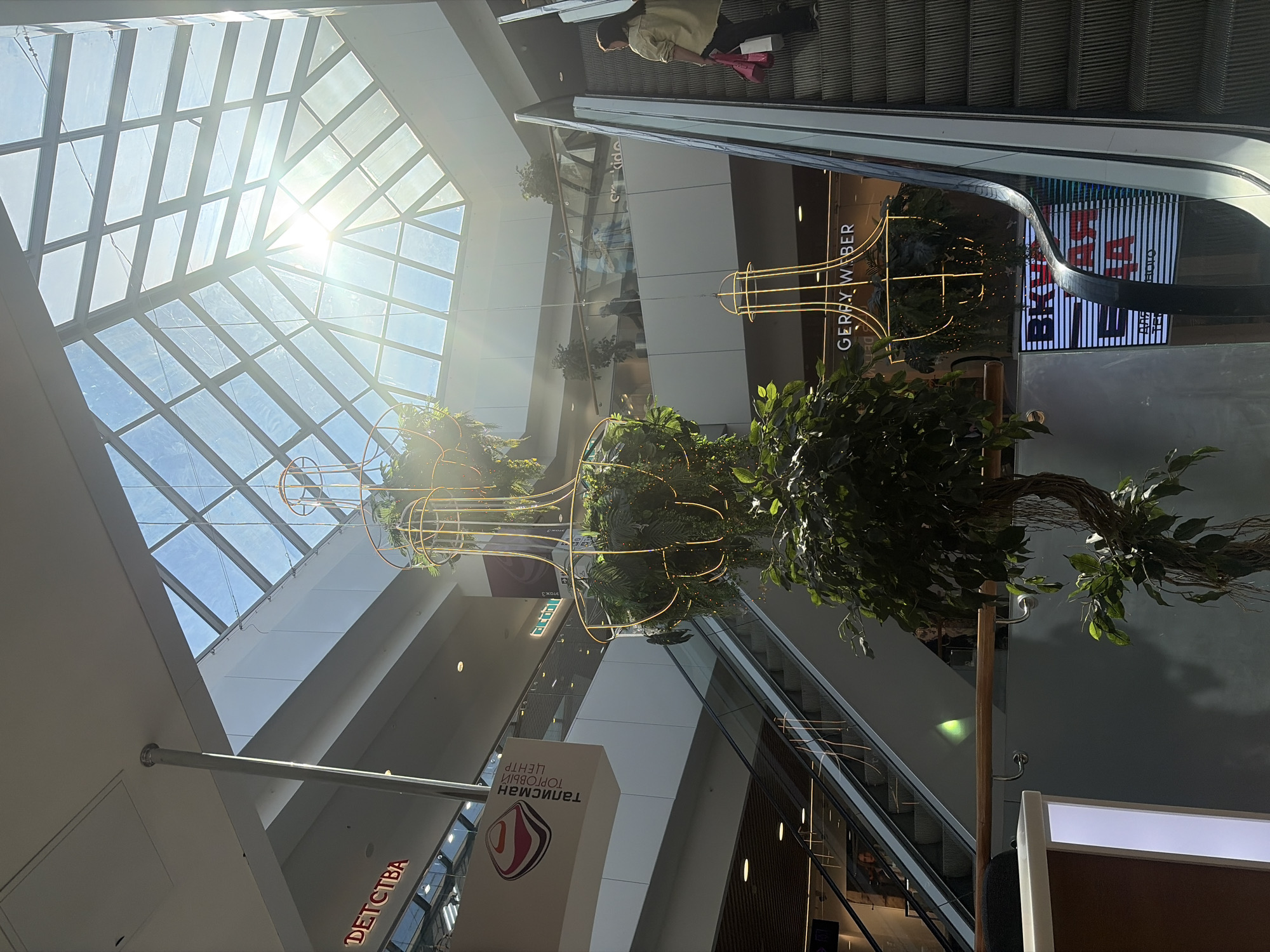 The image captures the vibrant interior of a multi-story shopping mall, likely the Talisman Shopping Center in Izhevsk, Russia, given the visible signage. The scene is brightly lit by natural sunlight streaming through a large, geometric glass skylight that forms the ceiling, creating significant sun glare in the upper left quadrant.
Multiple escalators are visible, connecting different levels of the mall. On the upper right, a person wearing a light-colored shirt and dark pants is ascending an escalator, carrying a pink bag. The mall features modern architecture with clean lines, white and grey walls, and glass railings on upper floors.
A prominent decorative element in the center of the mall is a multi-tiered plant display, consisting of artificial trees and greenery. Integrated within this display is a striking golden, cage-like sculptural structure, which appears to be illuminated or made of light-emitting material.
Visible text includes:
* "GERRY WEBER" on the facade of a clothing store.
* A large sign on a white pillar that reads "ТАЛИСМАН ТОРГОВЫЙ ЦЕНТР" (Talisman Shopping Center) in Cyrillic, accompanied by the center's abstract logo.
* Partially visible text "ДЕТСТВА" (Childhood), suggesting a children's store or section.
* Reflected text in shop windows, including "BARBARA" and "ETAGE 3" (Floor 3).
The overall impression is of a spacious and contemporary commercial environment, bustling with activity, taking place during a bright, sunny day.
