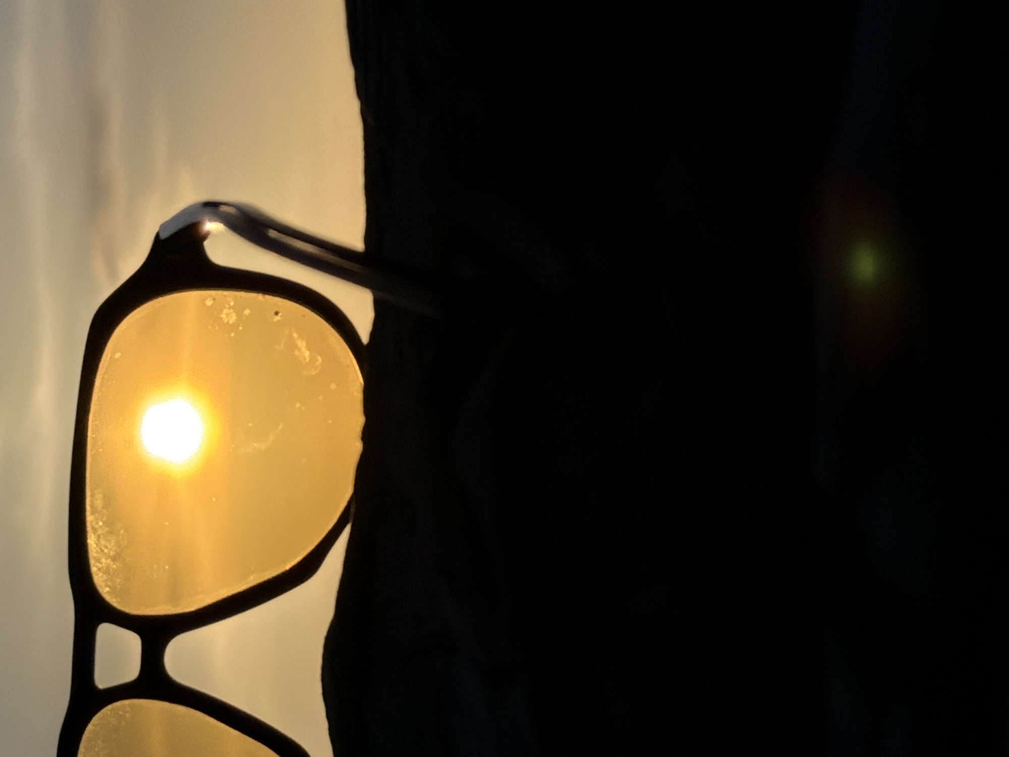 A pair of sunglasses is silhouetted against a golden sunset over Ko Samui District, Thailand. The lenses of the sunglasses reflect the bright sun, which appears as a luminous orb in the sky. The sunglasses are positioned in the foreground, with a dark, undulating landscape beneath them. The background consists of the hazy sky, with clouds tinged by the warm light of the setting sun. The overall impression is one of serene beauty, capturing a moment of tranquility as the day draws to a close.