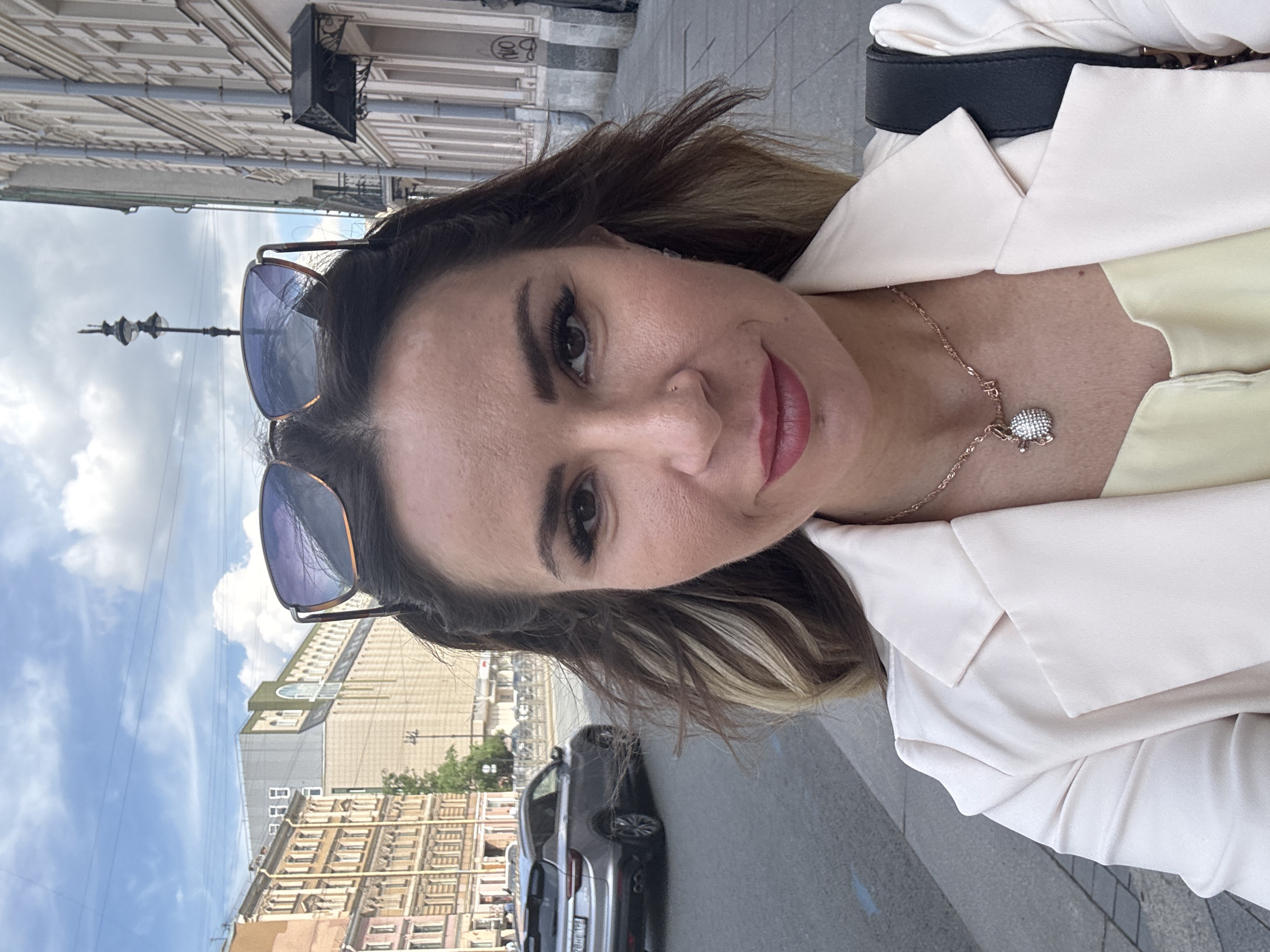 A woman stands on a sidewalk in Saint Petersburg, Russia, taking a selfie outdoors on a cloudy day. She is wearing a light-colored blazer over a yellow top and a necklace with a circular, diamond-encrusted pendant. Her sunglasses are perched on top of her head. In the background, a silver SUV is driving down the street, and several buildings with traditional architecture line the sidewalk. A lamppost is visible on the right side of the frame. The woman's hair is partially covering her face, and she is smiling.