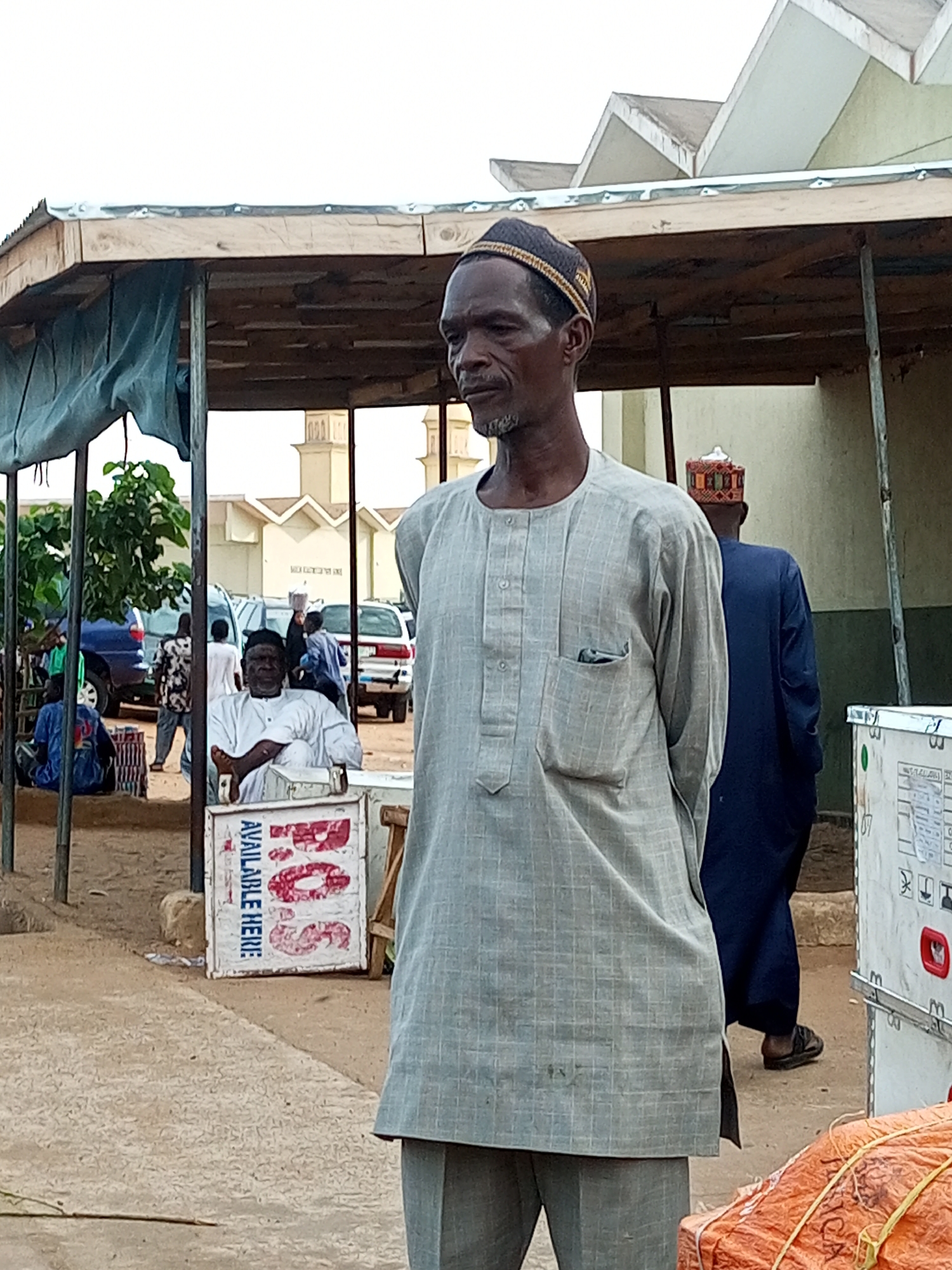 In Gombe, Nigeria, a man in a light gray tunic and cap stands looking to the side. He is under a wooden shelter with a corrugated metal roof. Behind him, another man in a dark blue tunic and a patterned cap walks away. In the background, there are vehicles, including a blue SUV and a white car. A mosque with two minarets is visible in the distance. A sign with red lettering and the words "AVAILABLE HERE" is leaning against a structure to the left of the man in the gray tunic. In the foreground, an orange-wrapped package is tied with yellow rope.