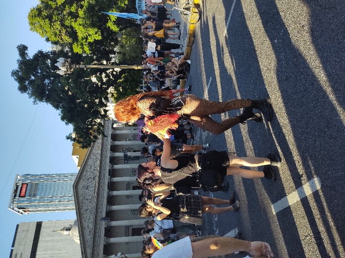 A street gathering features numerous individuals, including two distinctive figures: one with red hair and fishnet stockings, and another with a darker outfit. People are walking on the road in front of tall building with the sign “Galicia” on it. Some carry signs and wave flags. Sunlight casts shadows across the asphalt. A building with Roman columns is visible in the background. Trees and a lamppost stand along the side of the street.