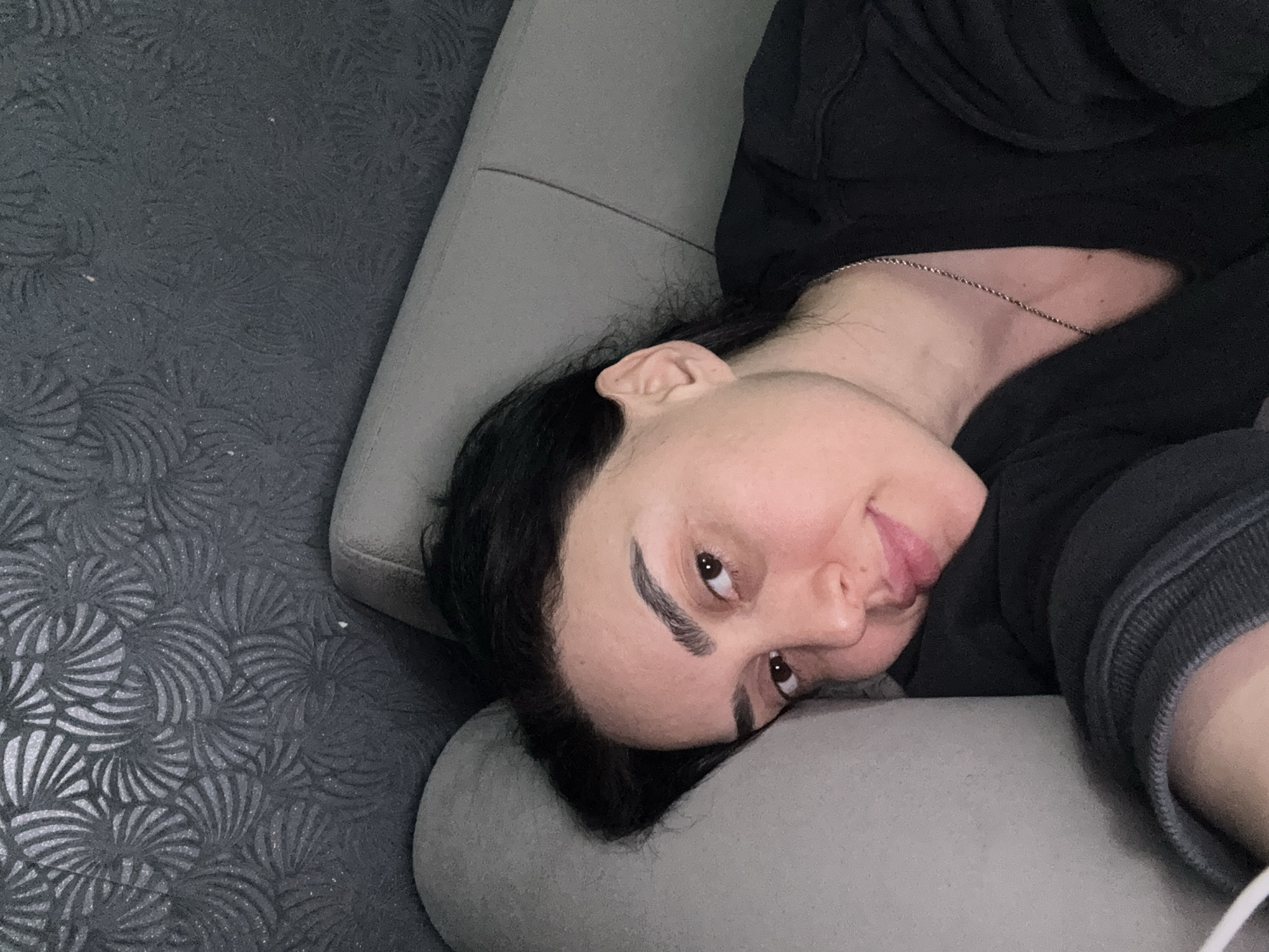 A close-up, diagonal shot captures a person, likely female, resting comfortably in what appears to be an indoor setting or vehicle in Tbilisi, Georgia. The individual is lying on their side, with their head and upper body supported by a light gray upholstered surface, possibly a sofa or car seat. Their dark hair frames their face, and they are looking towards the camera with a subtle, calm expression, featuring a slight smile or smirk.
The person is wearing a dark, possibly black, long-sleeved top or hoodie, and a thin silver-colored chain necklace is visible around their neck. Their features include dark eyes and prominent, well-groomed eyebrows.
In the background, to the left and upwards, a dark gray surface with a distinct, intricate pattern of swirling, fan-like, or stylized floral motifs provides a textured visual element, suggesting a carpet, rug, or specialized upholstery. A plain light gray upholstered surface runs vertically behind the person, serving as a boundary between the patterned background and the person. The lighting is soft and ambient, suggesting an indoor environment. The image conveys a sense of relaxation and casualness. No text is visible in the media file.