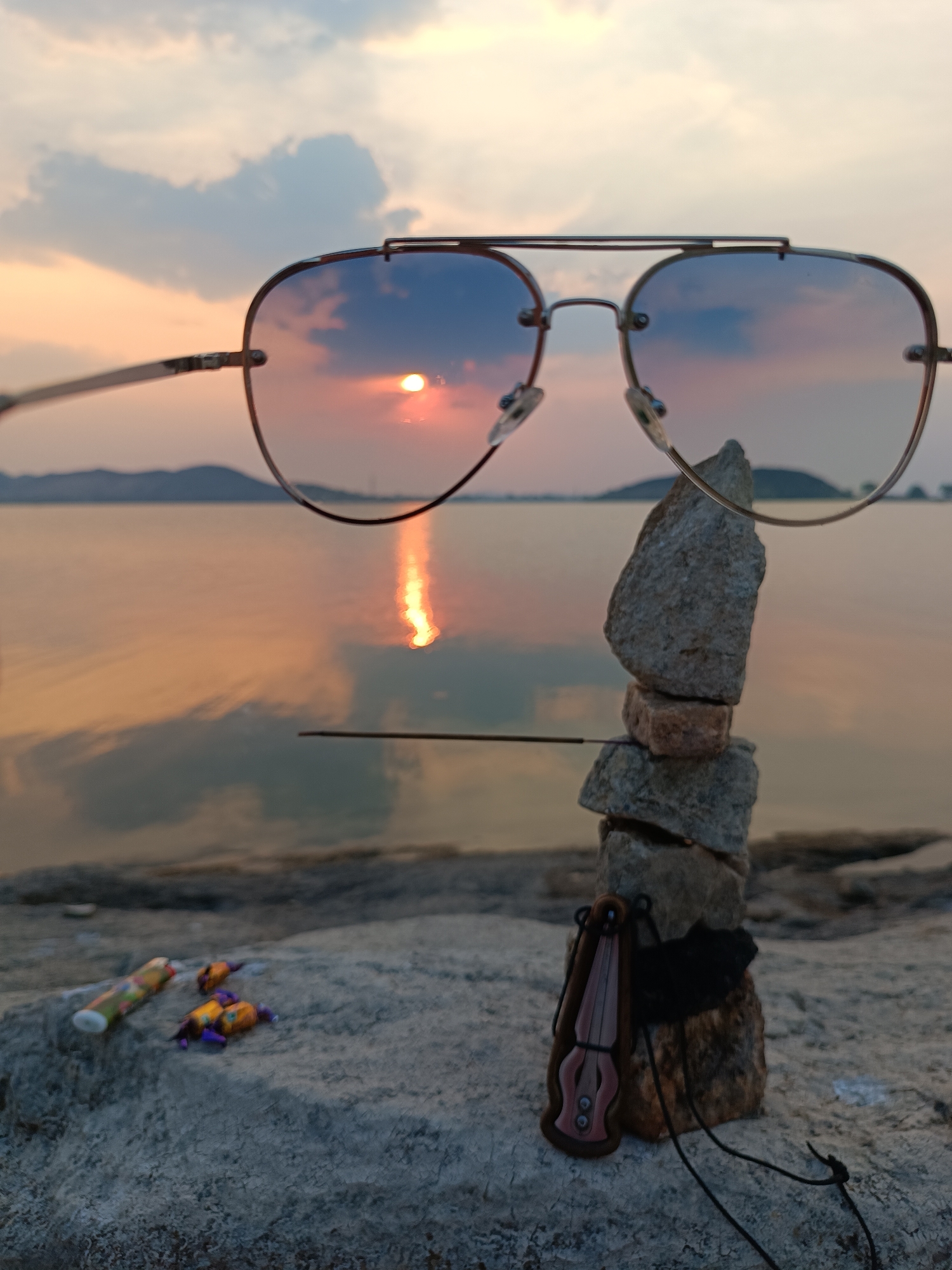 The image shows a stack of rocks with sunglasses perched on top, set against a backdrop of a body of water reflecting a sunset or sunrise. An incense stick rests against the rocks. Several wrapped candies are on the ground next to the stack. A wooden pendant or amulet is attached to the base of the rocks. The location appears to be near the water's edge. The city is Unknown, Unknown.