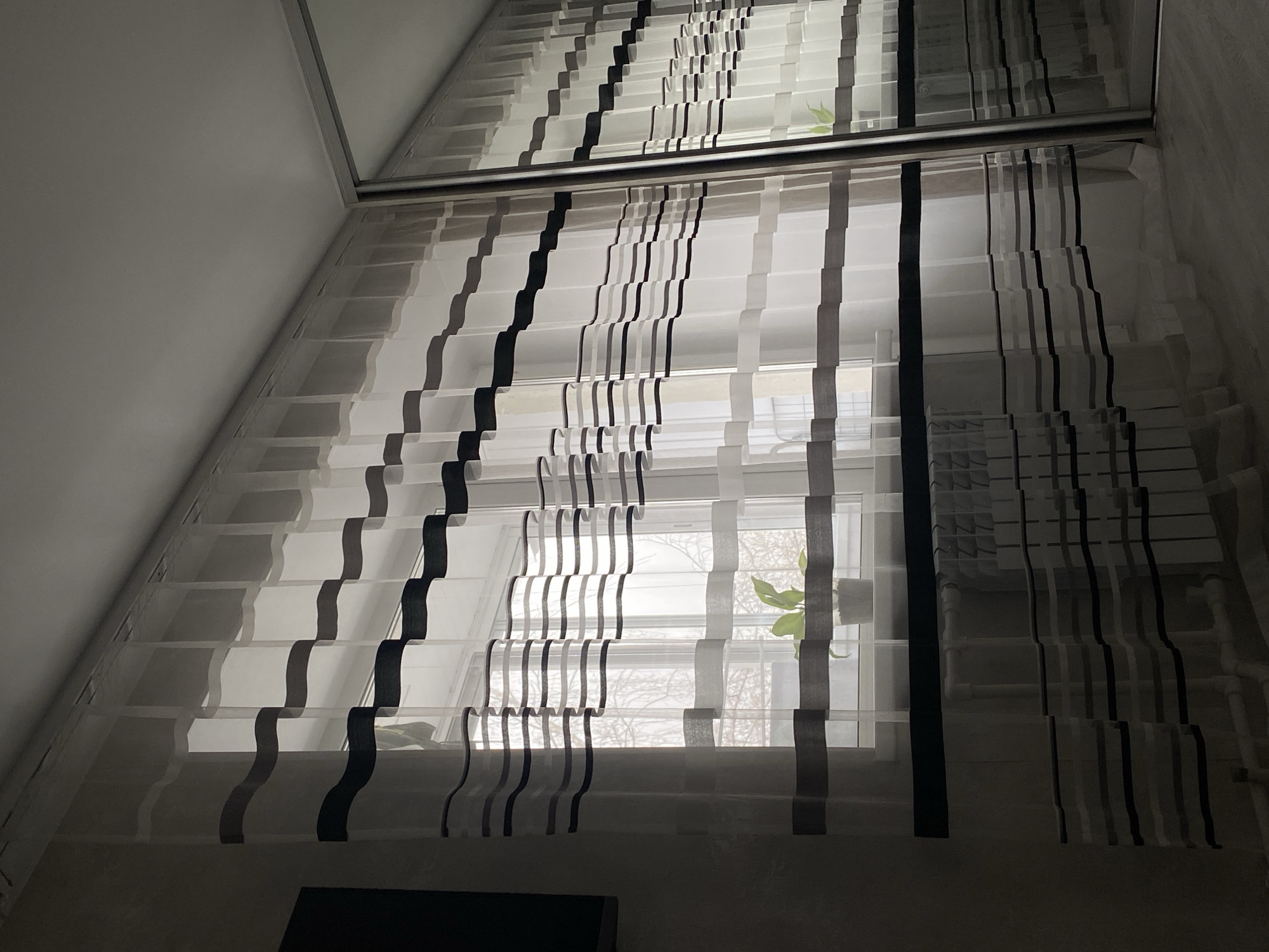 The image presents an interior scene, looking upwards at a large window fitted with patterned blinds or curtains. The perspective is tilted, with a light-colored wall or ceiling visible to the upper left and a textured wall to the upper right.
The primary subject is a set of sheer blinds featuring alternating horizontal strips of white/translucent, light gray, and dark gray/black fabric. These strips are arranged in a distinctive wavy, undulating pattern as they hang across the window. Through the translucent sections, a faint view of the outdoors can be seen, including what appear to be bare tree branches, suggesting a winter or early spring setting. A small green plant with a few leaves is visible near the window sill, partially obscured by the blinds.
Above the main window area, a dark horizontal frame separates it from a reflective surface, likely a mirror or glass panel, which strongly reflects the upper portion of the blinds and the room's ceiling. This reflection creates a visual doubling of the patterned fabric.
The lighting is soft and diffused, indicating daytime, possibly an overcast day, with no harsh shadows. No people or visible text are present in the image. The scene conveys a quiet, domestic atmosphere in Chaikovsky, Russia.
