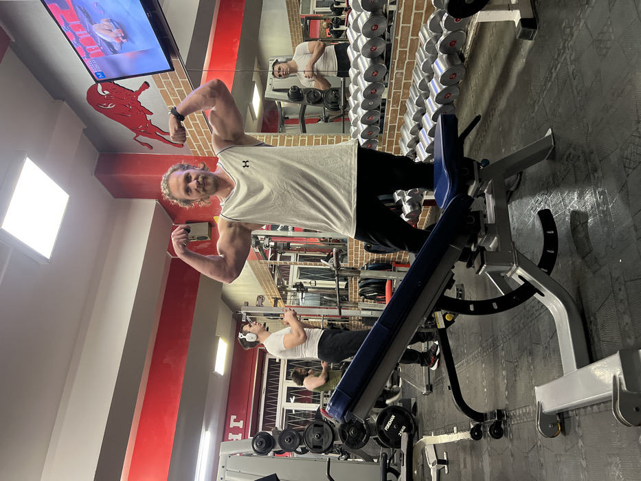 The image depicts an interior view of a commercial gym located in Saint Petersburg, Russia. The central figure is a Caucasian male, appearing to be in his 30s or 40s, with light brown curly hair and a beard. He is wearing a light grey sleeveless tank top with an Under Armour logo and dark trousers. He stands facing the camera, flexing both arms to display his biceps, with a subtle smile or smirk. A black wristwatch is visible on his left wrist.
In the foreground, an adjustable blue and grey weight bench is positioned in front of the main subject. The gym floor is covered with dark, interlocking rubber mats. To the left, in the mid-ground, another Caucasian male with short dark hair, wearing a white t-shirt and white headphones, is visible standing near a weight machine and appears to be interacting with a mobile phone. A third individual, partially obscured by equipment, is visible further left in a dark green top.
The gym features red and grey painted walls. A prominent red bull outline graphic is mounted on the upper right wall, adjacent to a flat-screen television displaying content labeled "Muscle Fitness." The right wall is extensively mirrored, reflecting rows of chrome dumbbells on a rack and other gym equipment. Various weight machines and barbell racks, including one with a "FOREMAN 25KG" weight plate, are visible on the left side of the frame. The ceiling is white with rectangular LED light panels providing illumination.