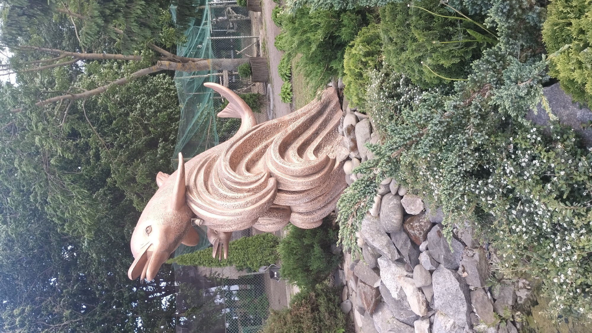 The image features a large, stylized sculpture of a dolphin, made of what appears to be bronze or a bronze-colored material. The sculpture is prominently displayed in an outdoor setting, possibly a park or garden. The dolphin's body is rendered with flowing, wave-like lines, suggesting movement and the fluidity of water.
The sculpture is positioned amongst natural elements. A rockery of grey stones forms a base for the sculpture and provides a transition into the surrounding greenery, consisting of various bushes and trees. The foliage includes evergreen shrubs and flowering plants with small white blossoms. The background features a fence and trees.
Based on visual evidence, the location is likely in a public or semi-public space, such as a park or garden. The city of Khmilnyk, Ukraine is provided as context, suggesting this sculpture may be a local landmark or artwork within that city.