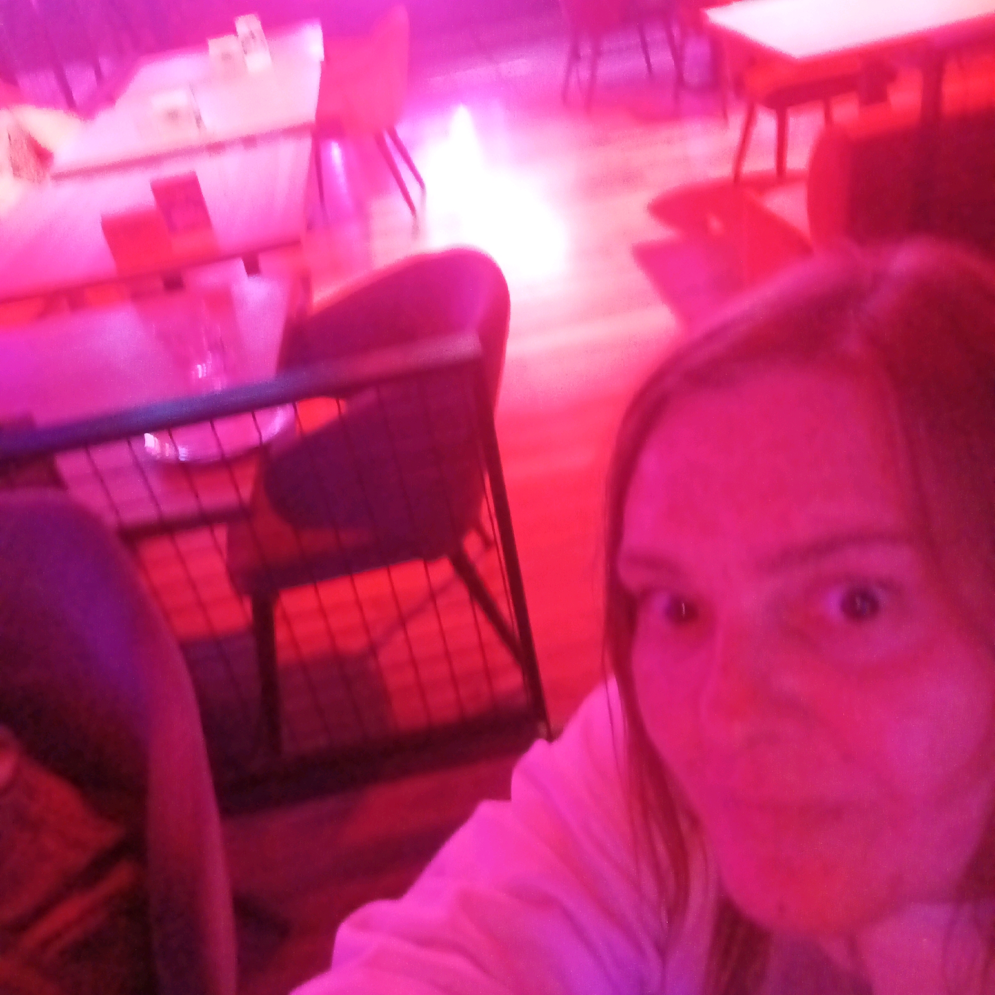 The image shows a close-up, slightly blurry selfie of a young woman with wide eyes and light-colored hair, bathed in intense pink and red light. She is wearing a light-colored top. The background appears to be an indoor setting with tables and chairs, suggesting a cafe, bar, or restaurant. The lighting creates a vibrant, possibly nightclub-like atmosphere. The image does not contain any information that would indicate the location is in Samara, Russia.