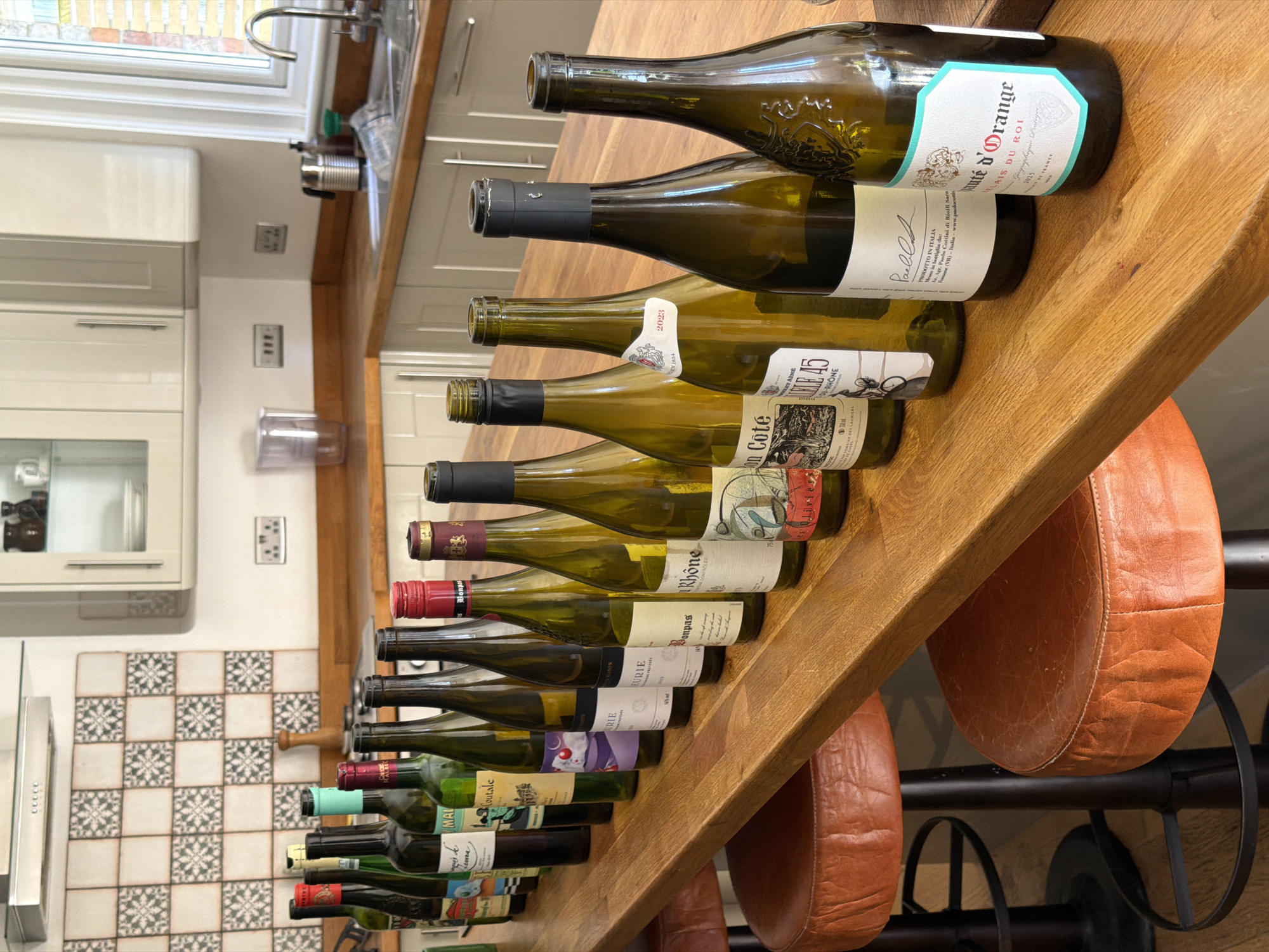 The image shows a collection of 17 wine bottles arranged horizontally on a wooden countertop. The bottles vary in shape and size, and their labels are partially visible. The labels indicate that some of the bottles contain wine from France and Italy. Brands such as "Mute d'Orange," "Paells," "Rhone," "Bonpas," "URIE," "Coté," and "LETAINE" are recognizable. The bottles are positioned in front of kitchen cabinets and countertops. The foreground includes two brown leather bar stools with black metal bases. The scene appears to be in a kitchen environment, possibly located in Folkestone and Hythe District, United Kingdom, though this cannot be confirmed from the visual data alone.