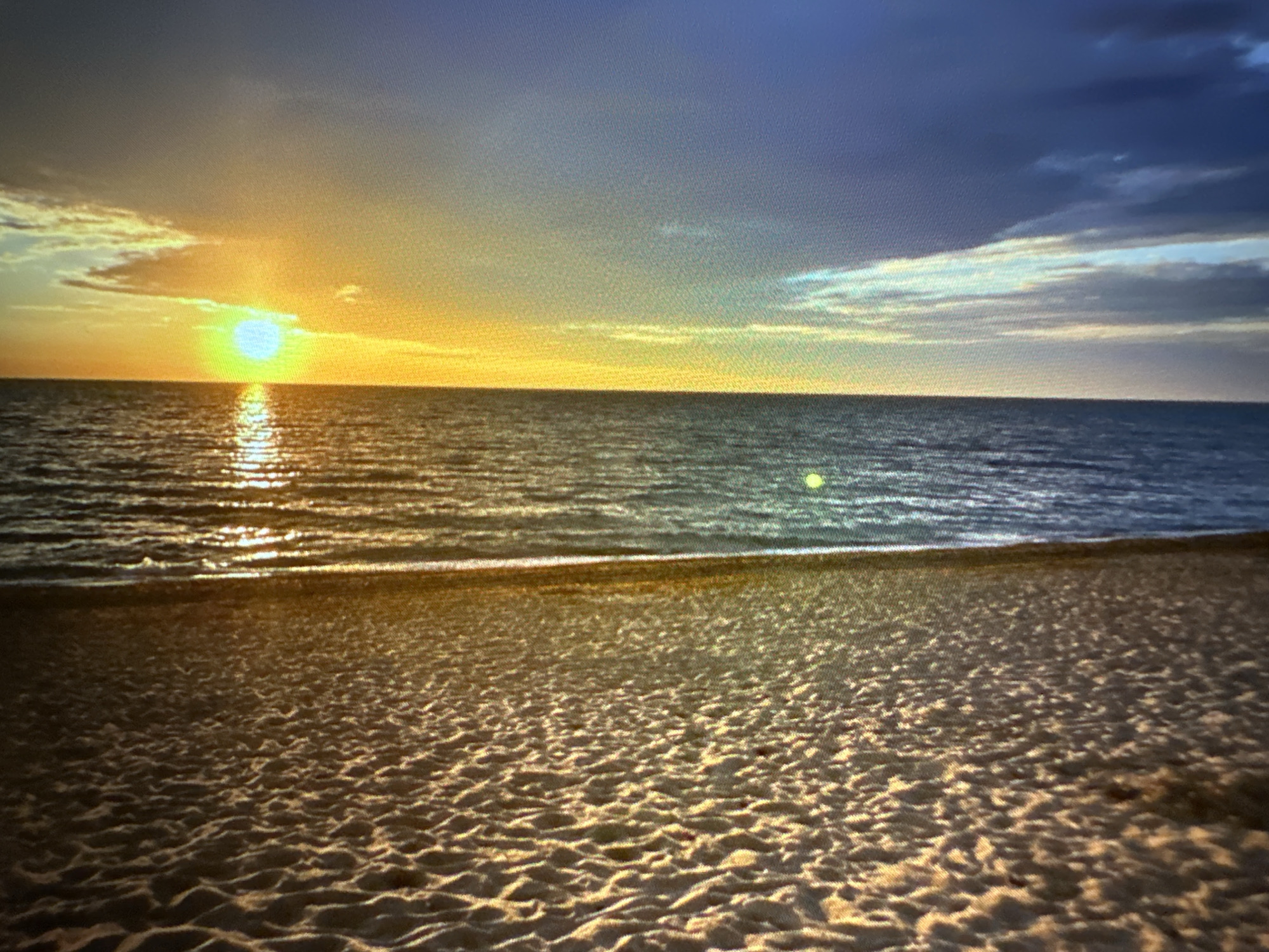 A vertically oriented, low-resolution image depicts a beach scene at sunset. The left third of the image is dominated by sand with a textured, rippled surface, suggesting it may be wet or have been recently disturbed. A thin vertical line separates the sand from the water. The water, occupying the middle third, appears dark and reflective, with vertical lines of light suggesting either ripples or the reflection of sunlight. The right third of the image is a vibrant display of the sunset. A bright, circular sun, emitting a greenish-yellow light, is positioned near the center of this section, casting a golden glow across the sky. Wispy clouds, tinged with orange and yellow, are visible in the sky, transitioning to a darker blue on the far right. A faint, greenish-yellow dot, possibly a lens flare or another light source, is visible in the water just to the left of the sun.