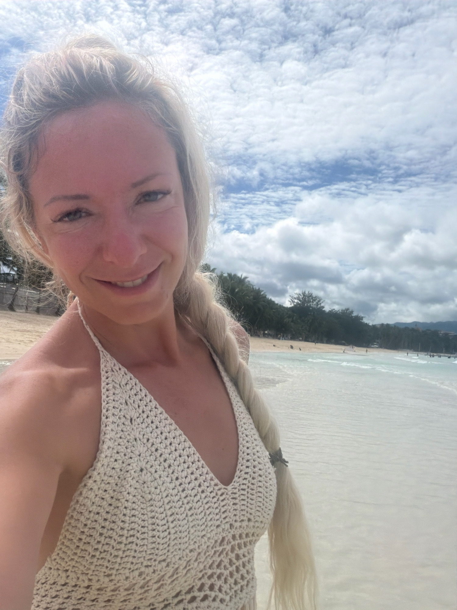 A woman with blonde hair braided down her back takes a selfie on a beach in Malay, Philippines. She is wearing a crocheted halter top and smiling at the camera. The beach is lined with palm trees, and the ocean is visible in the background with gentle waves. The sky is partly cloudy.