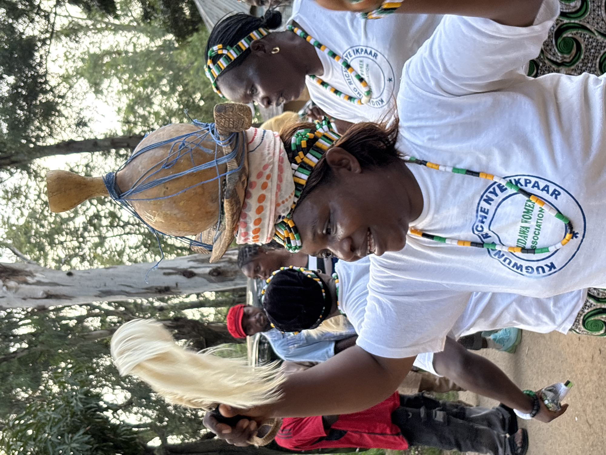 In a medium shot, a group of women are gathered outdoors, possibly in Jos, Nigeria, given the context. The foreground features a woman with dark skin, wearing a white t-shirt with a circular logo that reads "KUGWUNCHE NYE IKPAAR ASSOCIATIO N" and a colorful beaded necklace. Her right arm is raised, holding a bundle of white feathers. She is smiling, and her head is turned to her right. To her left, another woman, also wearing a white t-shirt and a similar beaded necklace and headband, is partially visible, looking to her left. Behind them, a woman with dark braided hair wears a black beanie and a colorful beaded headband. On her head, she balances a straw hat topped with fabric and a gourd secured with blue twine. In the background, a man in a red cap and light blue shirt looks towards the main subjects. To his left, a person in a red shirt is visible. The environment appears to be an outdoor setting with trees and foliage visible, and the ground is a dirt or gravel surface. The lighting suggests it might be late afternoon or early evening.