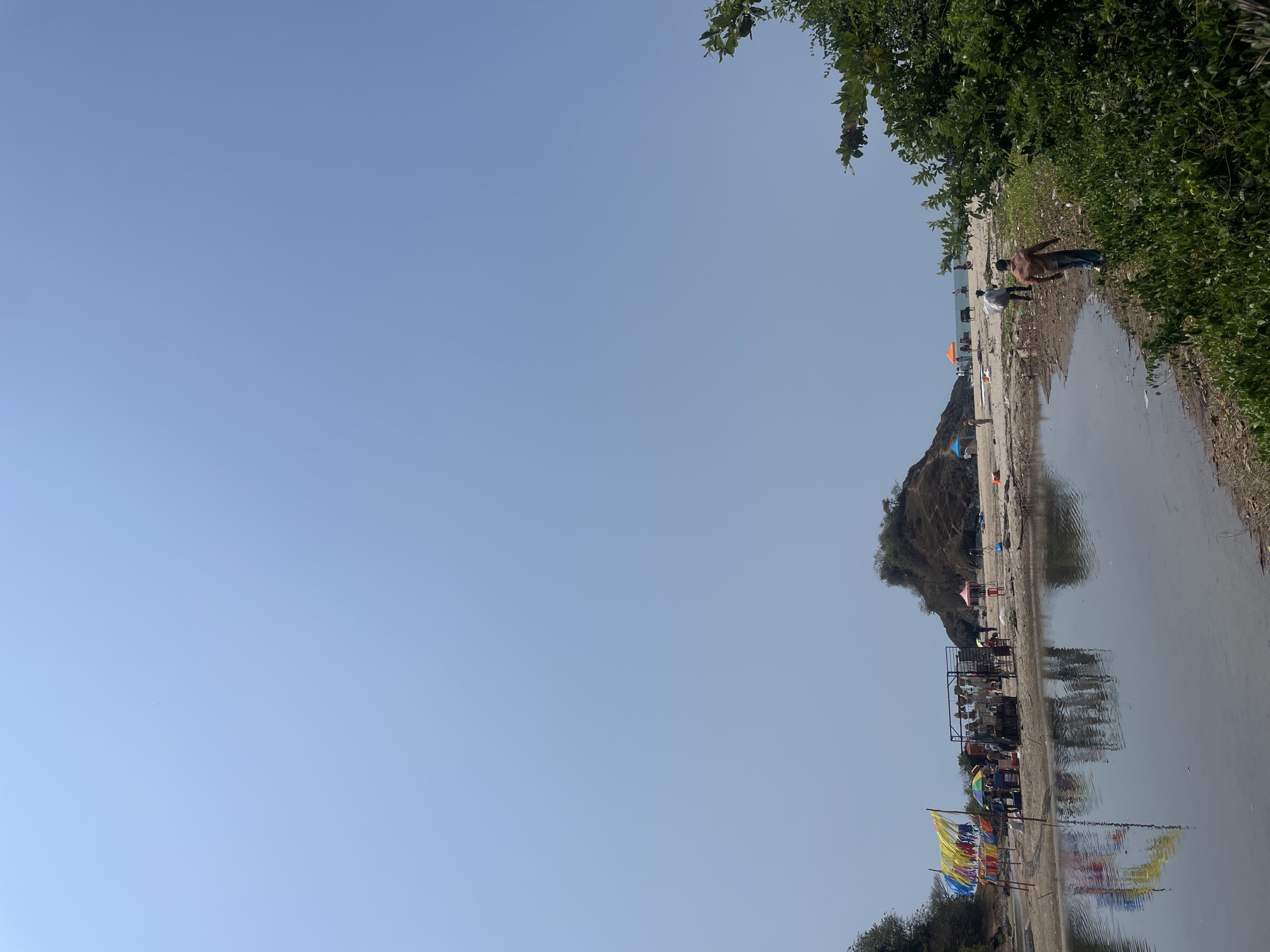 The image depicts a beach or riverbank scene in Bhogave, India. In the foreground, there is a body of water, likely a river or bay. A sandy shore stretches along the water's edge. People are visible along the shore. Structures resembling stalls or stands are present, along with colorful flags. A small hill or mound is in the background, possibly a natural land formation. The sky is clear. Vegetation is visible on the upper right.