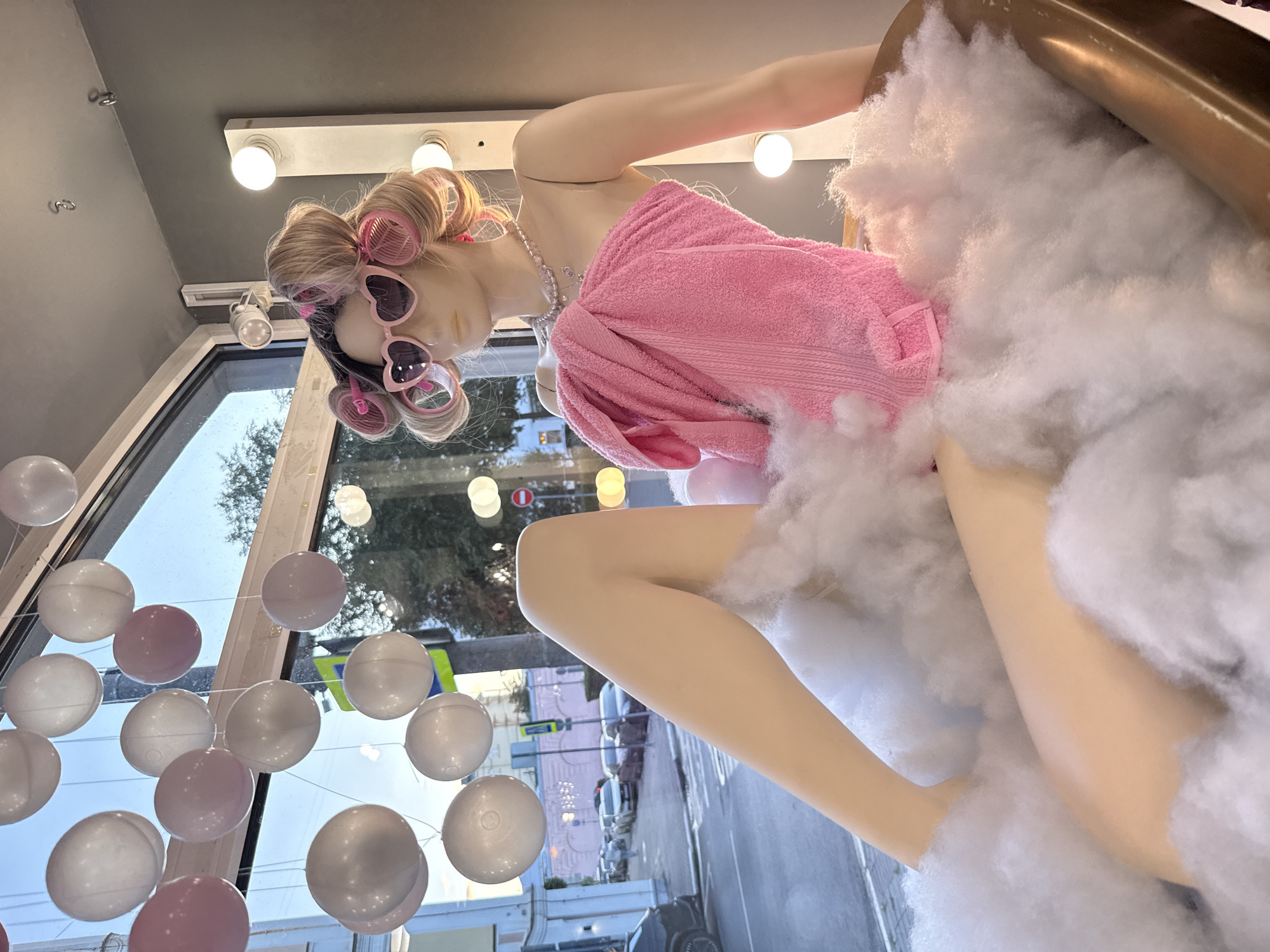 The image depicts a mannequin posed in a bathtub filled with fluffy white material resembling clouds. The mannequin has blonde hair styled with pink rollers, wears large pink heart-shaped sunglasses, and a pink towel wrapped around its torso. A pearl necklace is visible around its neck. In the background, through a large window, a street scene in Rusanovo, Russia, is visible, showing cars and buildings. The window is decorated with hanging white and pink circular ornaments. The scene appears to be a window display.