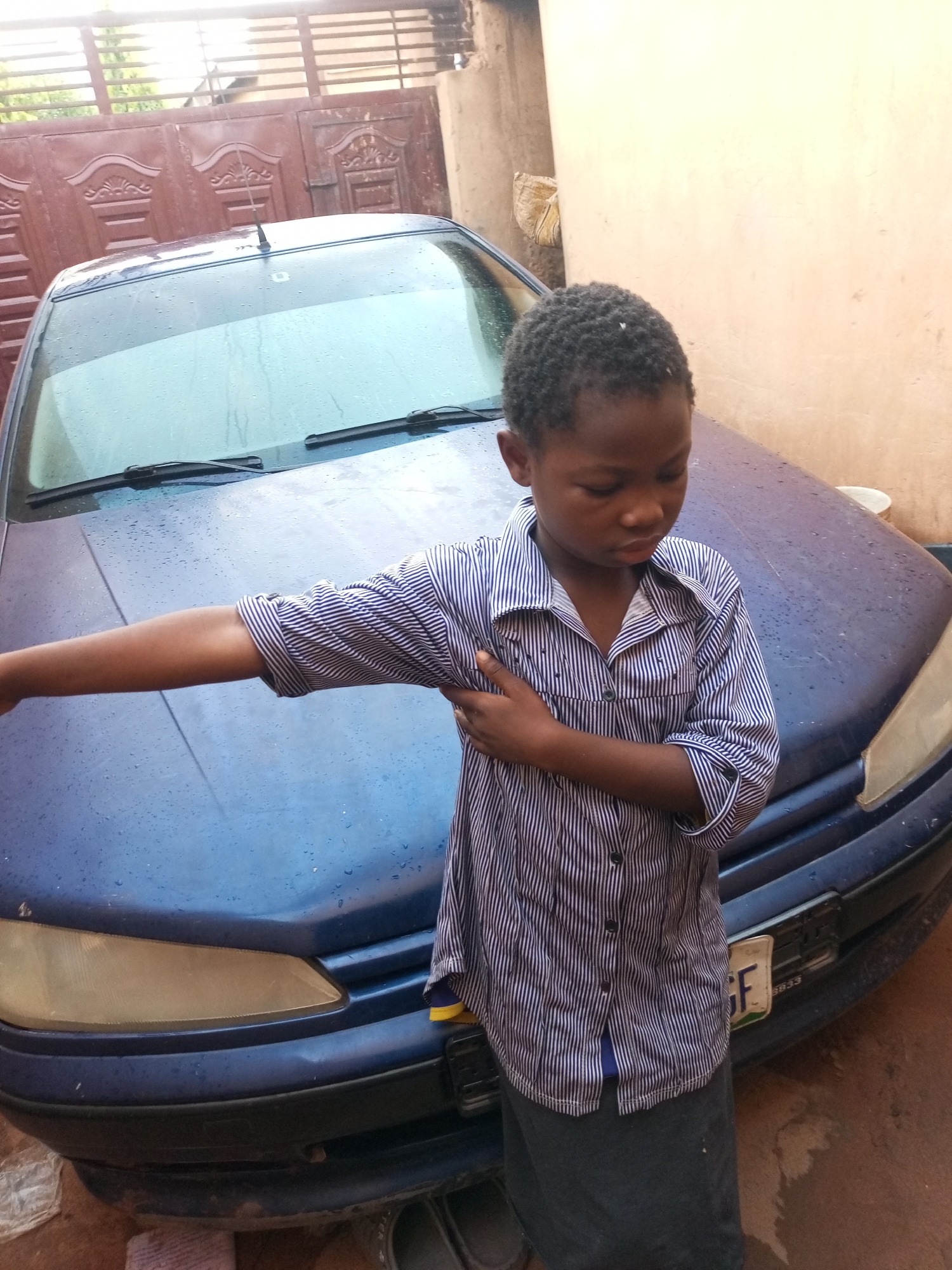 A young person, appearing to be a child, stands in front of a blue car. The child is wearing a striped button-down shirt and a dark skirt. Their right arm is bent and resting on their left arm. The car's hood is wet, indicating recent rain or washing. Behind the car, a large, ornate metal gate is visible, with a textured wall to the right of the child. The ground appears to be made of dirt or concrete and is also wet. The scene is likely outdoors, given the presence of natural light and what appears to be a residential or outdoor storage area.