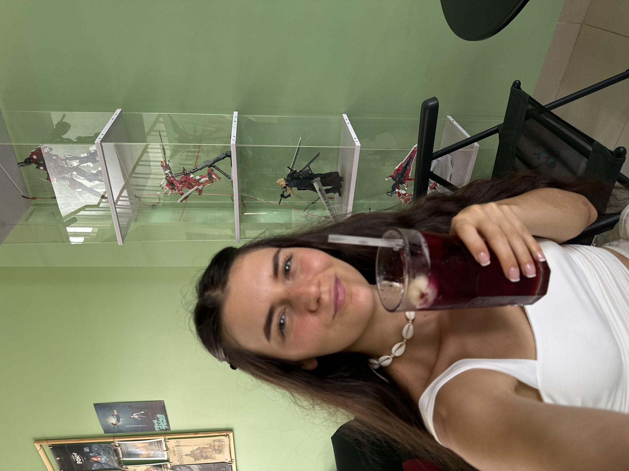 A young woman in Phu Thuy Ward, Vietnam, is holding a glass of a dark red beverage with ice and a straw. She wears a white top and a seashell necklace. Behind her is a green wall with five stacked, clear display cases containing model figures including mecha and a figure wielding swords. Below the display cases are two framed posters. A black folding chair is on the right side of the image. A black circular table is in the upper right corner.