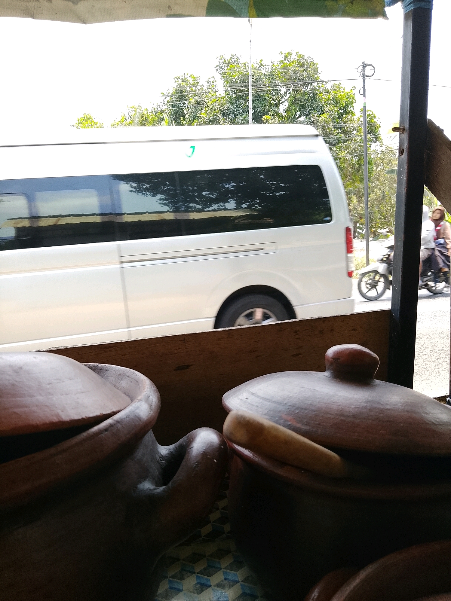 In Banaran, Indonesia, a white van is parked on a paved road. Trees line the street behind the van, and a utility pole stands to the right. A motorcycle with two people on it is riding past the van. In the foreground, two clay pots are visible, one on the left and one on the right. The pot on the left is dark brown and has a lid. The pot on the right is also dark brown and has a lighter brown lid. Both pots have handles.