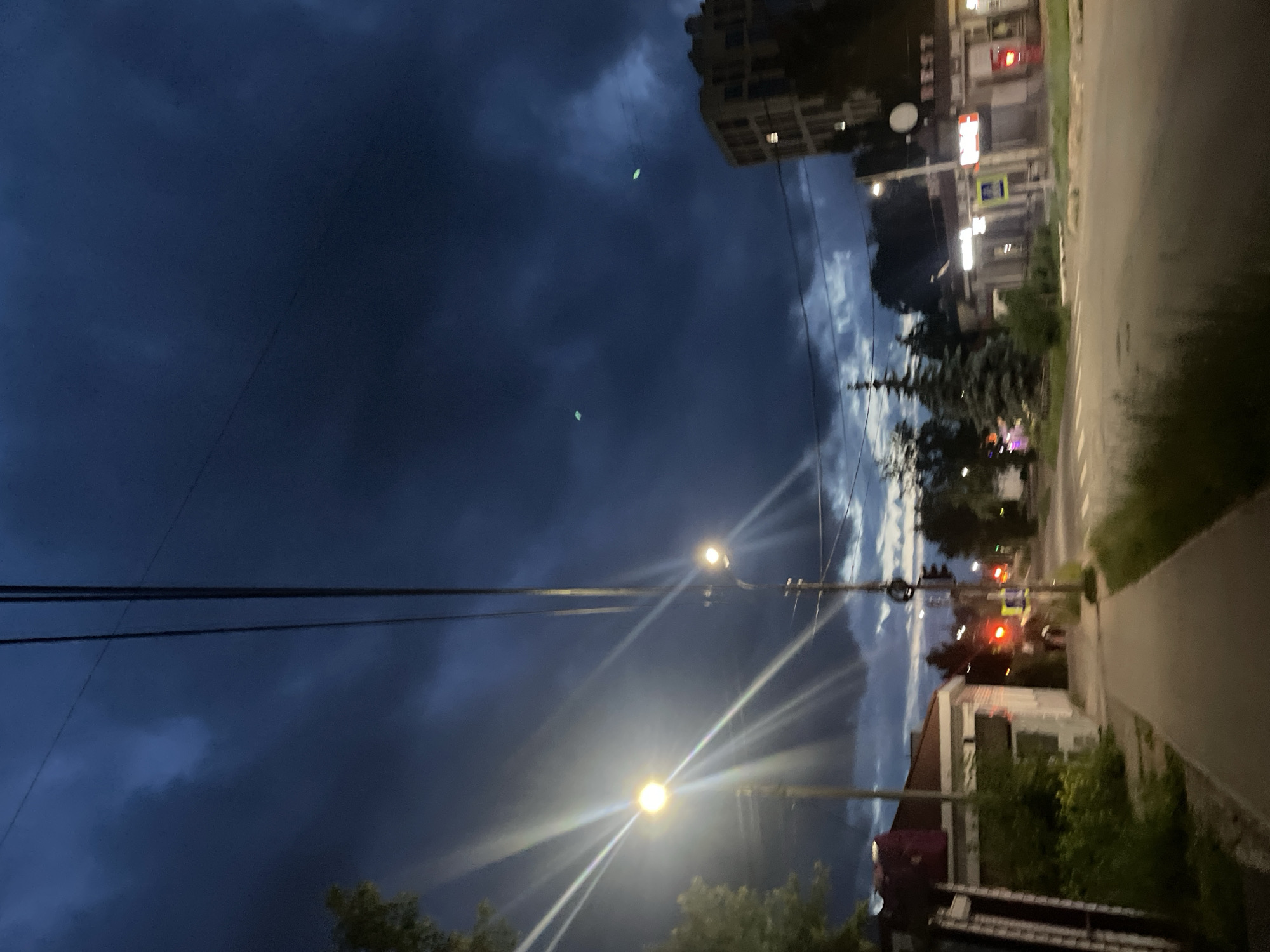 This media file depicts an outdoor scene at dusk or early evening. The sky is filled with dark, ominous clouds, suggesting impending rain or a storm. The overall mood is somewhat dramatic due to the lighting and cloud cover.
The scene appears to be a street or roadside in a residential or semi-commercial area, likely in Votkinsk, Russia, based on the provided context. Visible elements include power lines strung across the sky, with several streetlights illuminating the area. Some of these lights are bright and create lens flare effects, particularly the lower one.
In the background, there are buildings, one of which is a taller apartment-style building with visible windows, some illuminated. Other buildings appear to be single-story structures, possibly shops or houses. There are trees and vegetation scattered throughout the scene, some of which are silhouetted against the darker sky.
A road or paved pathway runs along the right side of the frame, with a grassy verge bordering it. Traffic lights are visible in the distance, showing red lights, indicating traffic is stopped. There are also other illuminated signs or shop displays, but the text is not clearly legible due to the low light conditions and image quality. There are no people clearly visible in the frame. The orientation of the image is such that it appears to have been taken looking upwards and sideways, possibly from a moving vehicle or handheld device.