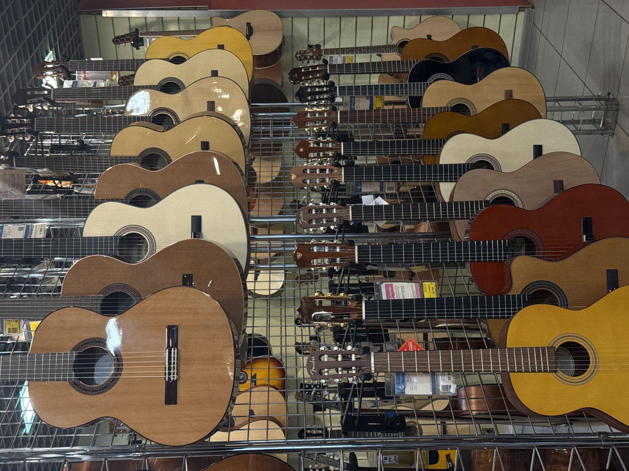 A store display in Moscow, Russia, presents numerous classical guitars. The guitars are arranged on a multi-tiered, wire-frame shelving unit. The instruments vary in color from light yellow to dark brown and red. Their headstocks and bodies are aligned, indicating orderly placement. Retail tags are visible amidst the instruments.