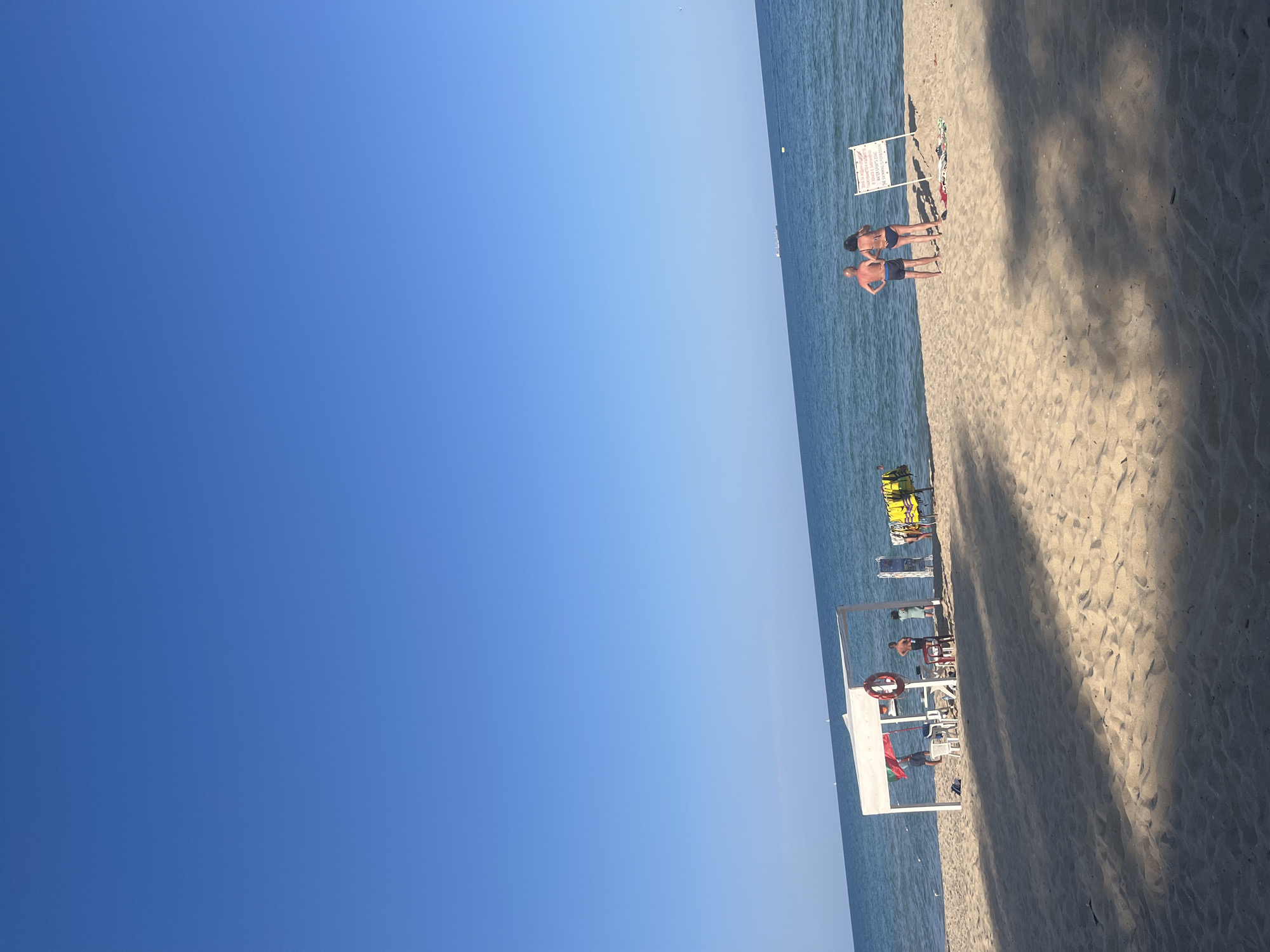This is a wide, eye-level shot of a sandy beach in Obzor, Bulgaria. The sky is a clear, bright blue, with the horizon line where the sky meets the calm, blue sea dominating the top half of the frame. In the middle ground, there is a white canopy structure with a table and chairs set up underneath it. A red life preserver is hanging from one of the canopy's supports. Several people are standing around and under the canopy, some appear to be staff members. To the right of the canopy, there are a couple of people in swimwear standing on the beach, facing the sea. A white sign with text is visible behind them. Yellow life jackets are hanging on a rack to the left of the sign. The sandy beach has some shadows cast across it, indicating it is a sunny day.