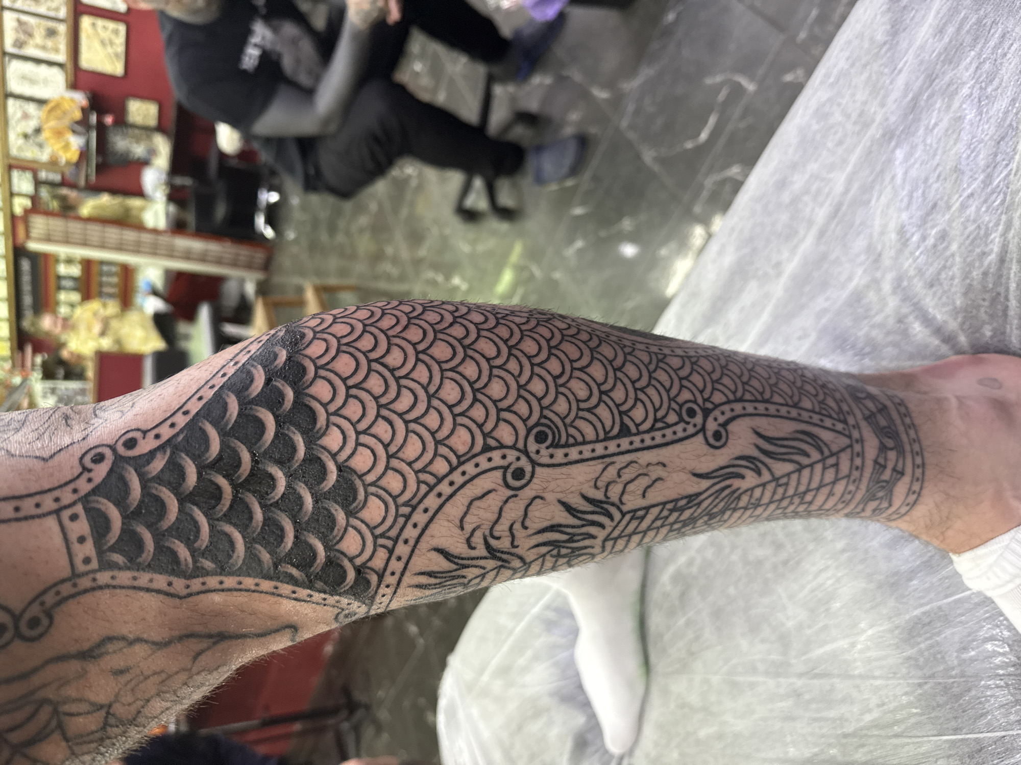 A close-up, high-angle shot shows a person's left forearm and lower leg, both covered in a detailed black-ink tattoo featuring a dragon-scale pattern, with a dragon head near the wrist. The arm is hairy, and the leg is resting on a white, possibly medical-grade, cover. In the blurred background, a person with tattooed arms is seated in a chair. The setting appears to be a tattoo studio, recognizable from the blurred images of artwork and equipment in the background.
There is no information to assume the location is in Moscow, Russia.