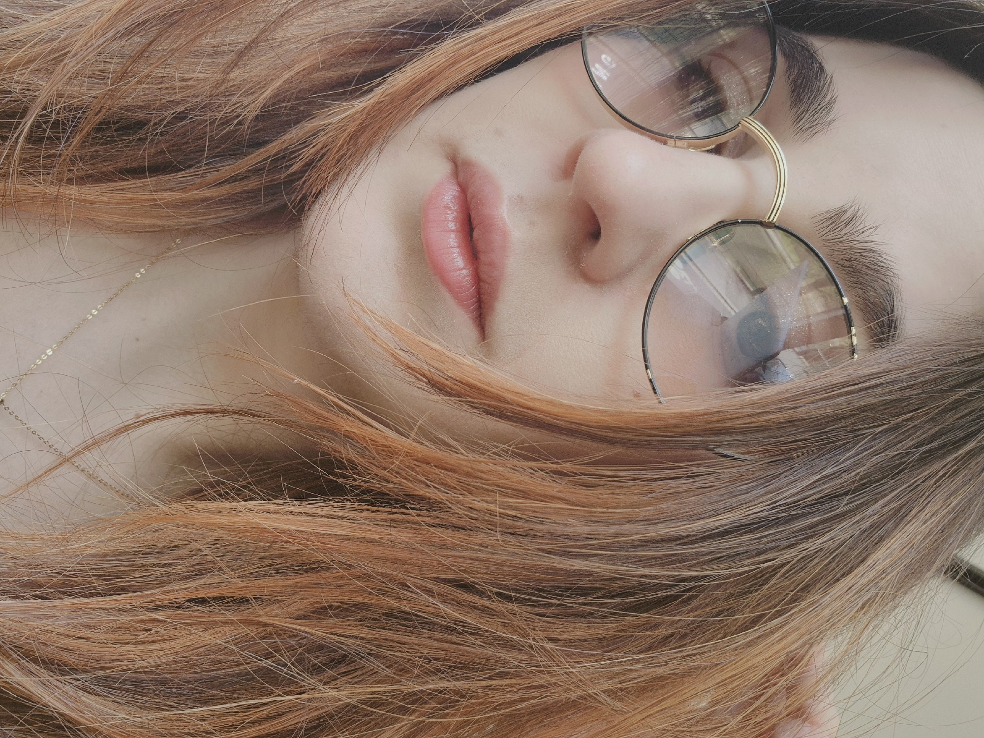 A close-up shot features a person with reddish-brown hair, lying at an angle. The focus is on their face, where they wear round, black-rimmed glasses with gold nose pieces. The person's lips are slightly parted, showing a pink hue. A thin gold necklace is visible along their neck. Due to the limited visual data, the location within Strasbourg, France, cannot be determined.