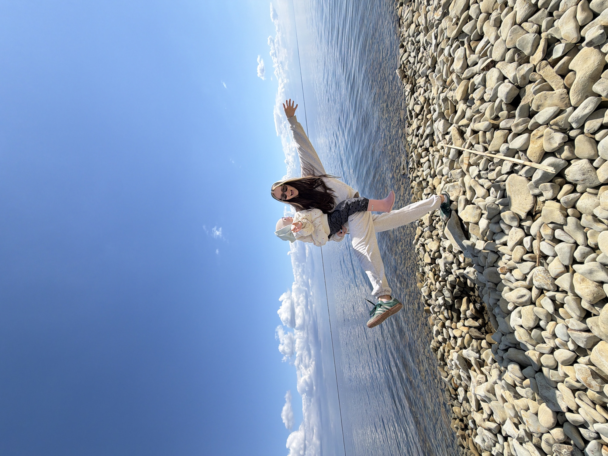 A woman in a light-colored tracksuit and sunglasses holds a child in her arms, raising her right arm. The child wears a light-colored outfit and pink boots. They are on a rocky beach next to water, with a clear blue sky with white clouds above. A bundle of sticks lies on the rocks. The location appears to be a waterfront area, possibly in Kamyshin, Russia.
