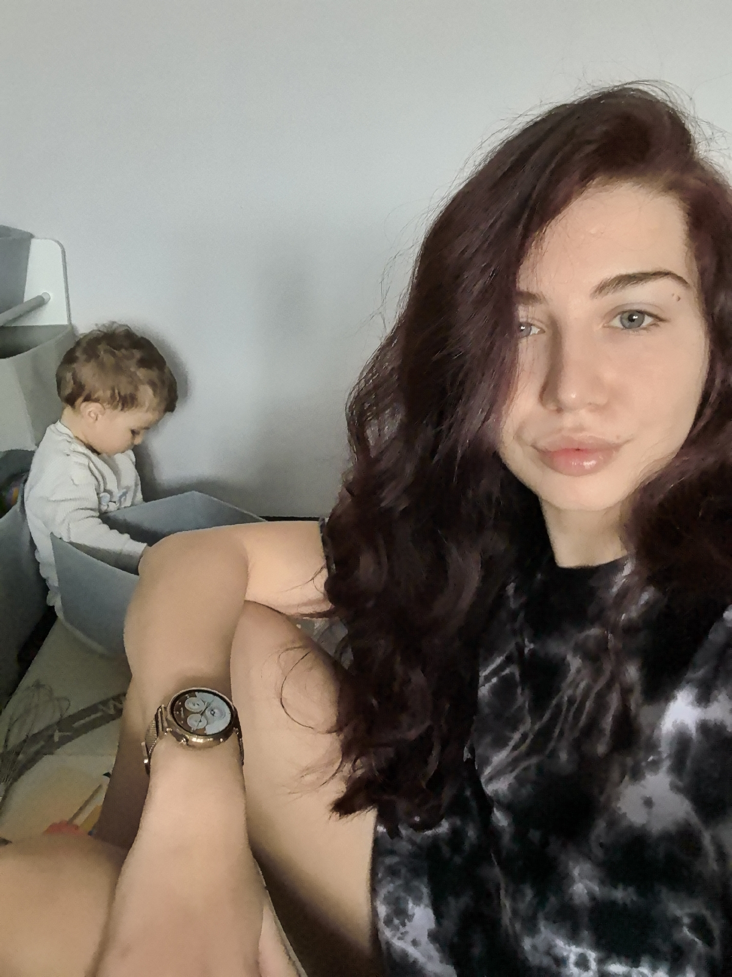 The image captures an indoor scene in Stara Zagora, Bulgaria, featuring two individuals. In the immediate foreground and to the right, a female subject, appearing to be an adult, is visible from the chest up, looking directly at the camera. She has long, dark reddish-brown wavy hair, light skin, and blue-green eyes, with full lips. She wears a black and white tie-dye patterned top. A gold-toned smartwatch with a circular face and a mesh band is present on her left wrist. Her bare legs are partially visible below the waist. In the mid-ground, to the left, a young child, approximately 1-3 years old, with light brown curly hair, is positioned within a grey fabric storage bin. The child is wearing a light grey long-sleeved shirt and is looking downwards, away from the camera, appearing to be focused on contents within the bin. The background consists of a plain, light-colored wall. No direct interaction is observed between the two individuals.