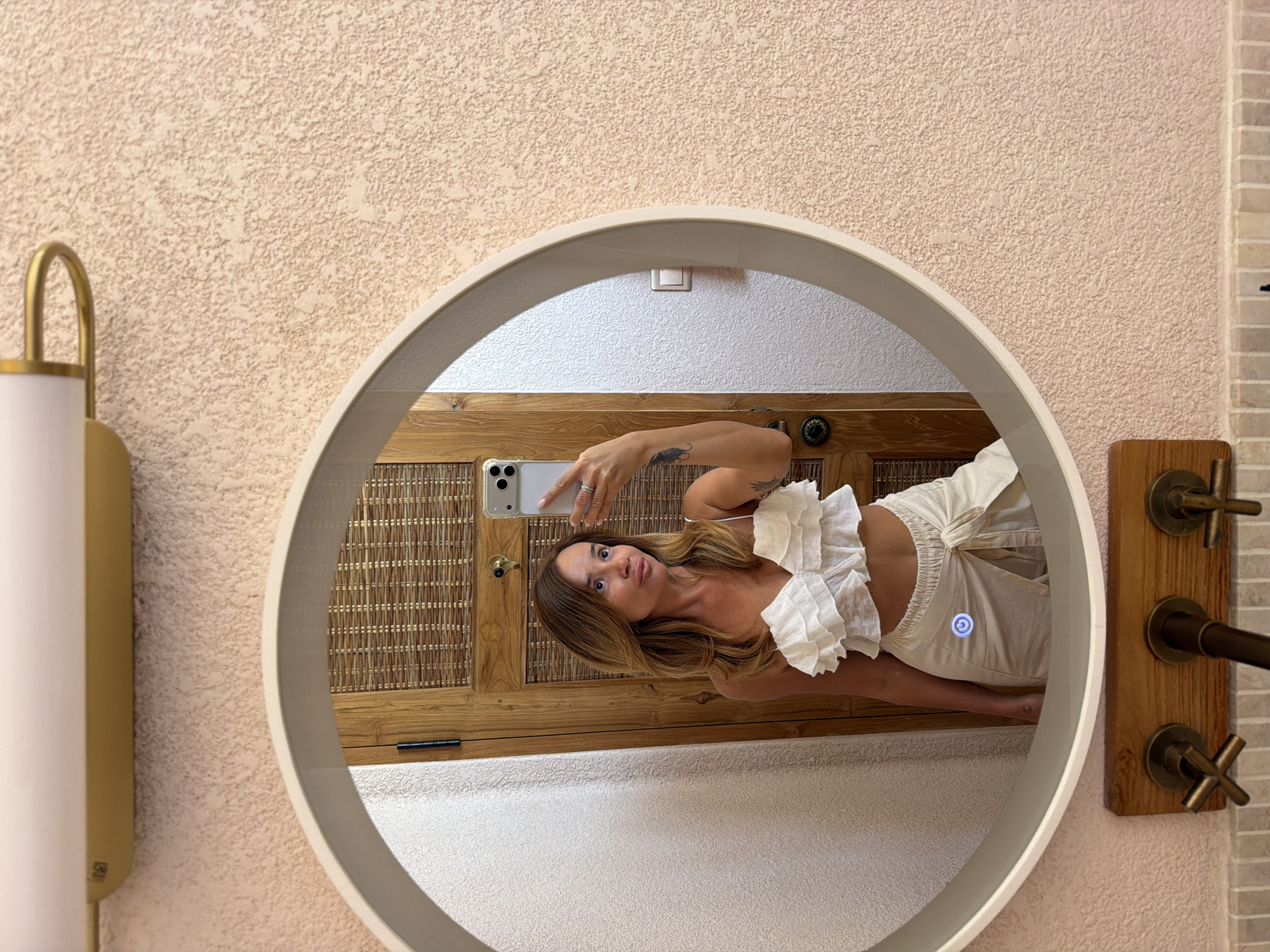 A woman stands in front of a textured pink wall, taking a mirror selfie. She is wearing a white ruffled crop top and beige wrap-around skirt. The mirror reflects a wooden door with a woven inlay. Below the mirror, a wooden fixture holds three cross-shaped faucets. The lighting fixture above the mirror is gold and modern. The scene appears to be in a well-lit bathroom.