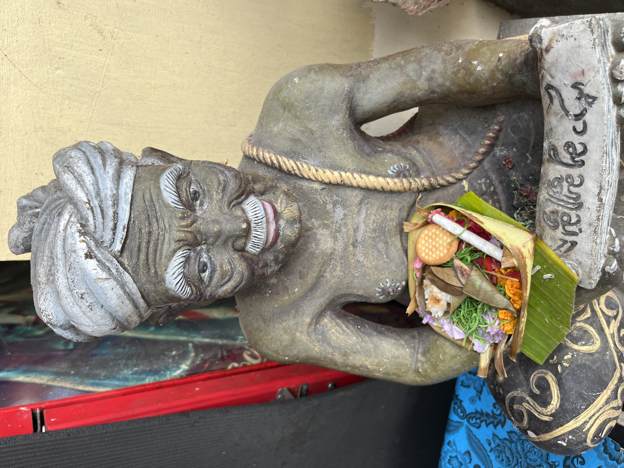 The image shows a weathered statue of an elderly man wearing a turban and a rope across his chest. The statue is holding a small offering in a banana leaf basket. The offering contains rice, flowers, a biscuit, and some unidentifiable items. The statue is seated, and its base has what appears to be writing on it. The statue is placed outdoors, with a yellow wall in the background. To the left of the statue, there is a red object and a black textured surface. The overall appearance suggests it is a religious or cultural artifact, possibly located at a temple or shrine in Kuta Selatan, Indonesia.