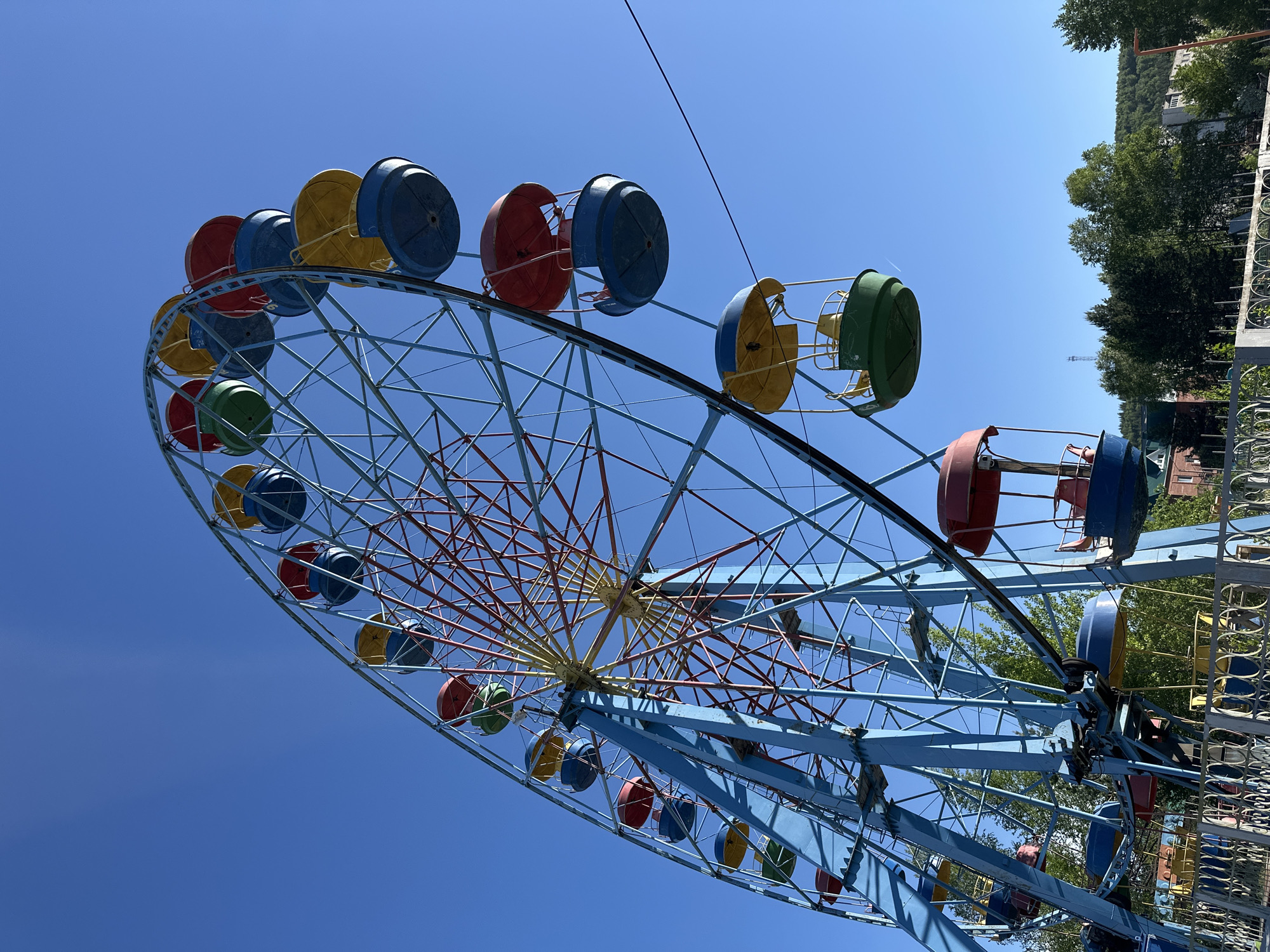 The image shows a Ferris wheel in the city of Unknown, Unknown. The wheel has a blue frame with red and yellow accents in the center. The passenger cars are individually colored in red, blue, yellow, and green. The background consists of a clear blue sky and some trees on the right side of the frame. Some buildings can be seen behind the trees. In the lower right corner, part of a fence is visible.