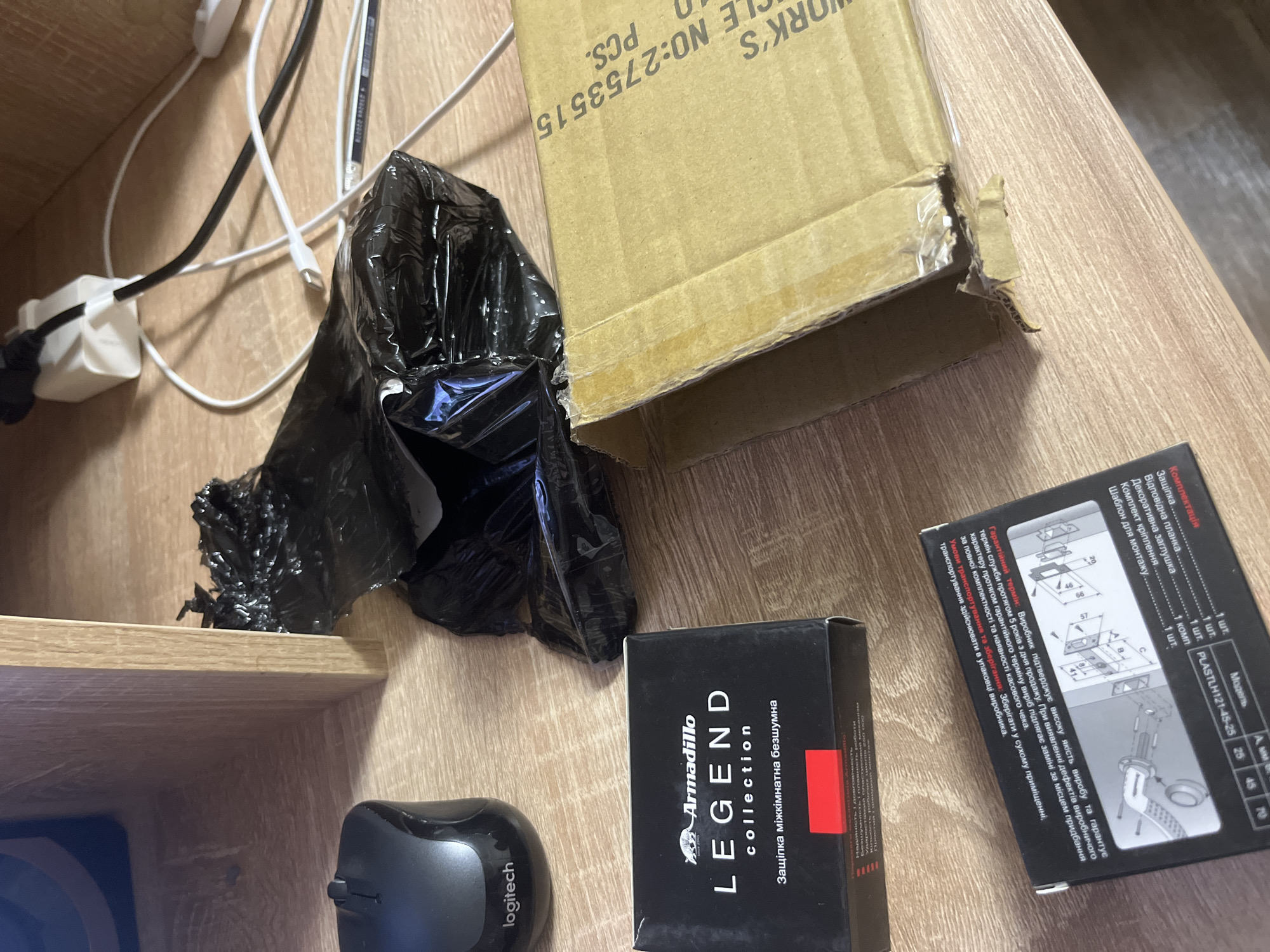 The scene depicts a table or desk surface in an indoor environment, likely a home, as indicated by the personal items scattered around. A black Logitech wireless mouse is positioned towards the bottom left corner of the frame. Next to it is a small black box labeled "Armadillo LEGEND collection," featuring Ukrainian text that translates to "Silent Interior Latch." Adjacent to the box is another package, bearing diagrams and details about the product, including its model (PLASTLH121-45-25) and dimensions (25, 45, 70). Above these items, there's a black plastic bag, crumpled and partially open. A partially visible cardboard box is located near the upper right area of the frame, with the text "WORK'S" and a serial number "2753515" visible. In the upper left quadrant, there's a shelf edge and visible electrical cords and a white power adapter. The table surface has a light brown wood grain pattern. The scene appears disorganized, with several unrelated objects placed randomly on the surface.