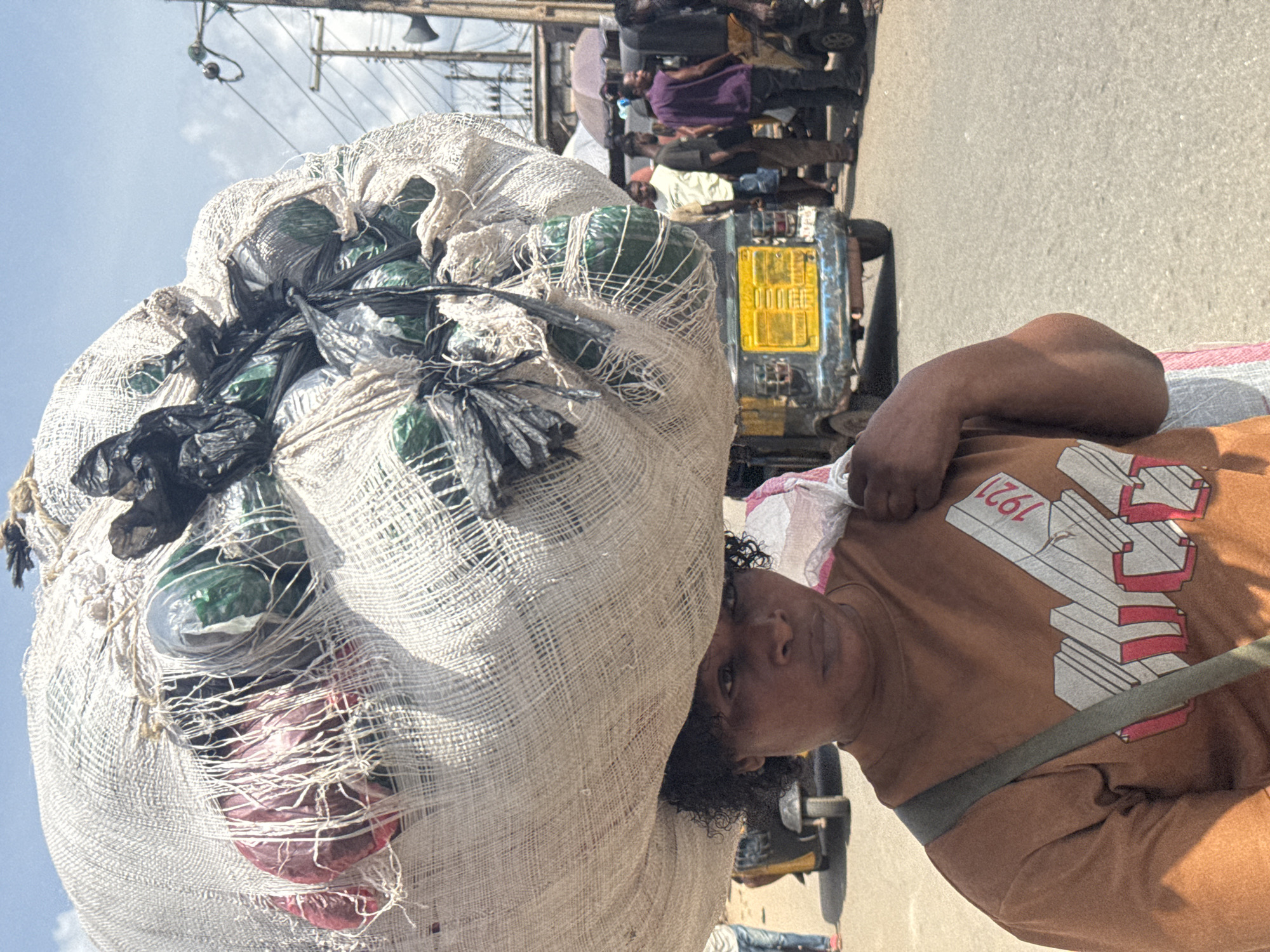 The image captures a vibrant street scene in Onitsha, Nigeria, centered on an individual carrying a large, heavy bundle on their head. The main subject, appearing to be a woman, is positioned towards the right of the frame, looking directly at the viewer with a neutral expression. She wears a brown short-sleeved t-shirt featuring a prominent design with the numbers "1921" in white and red, rendered in a three-dimensional style. A dark strap crosses her chest, possibly from a bag or additional carrying apparatus.
The significant load balanced on her head is encased in a coarse, light-colored mesh fabric, likely jute or a similar material. Through the netting, various items are visible, including multiple plastic bottles and other goods wrapped in black and green plastic bags, suggesting a cargo of recyclables or merchandise.
The background reveals a bustling urban environment. Other people are partially visible, some riding or standing near vehicles. A yellow license plate, characteristic of a commercial tricycle (like a Keke Napep common in Nigeria), is visible behind the main subject's head, though specific characters are largely obscured. Utility poles and overhead wires crisscross against a bright, clear blue sky, indicating an outdoor setting during the day. Strong sunlight suggests it is likely midday or early afternoon. The overall scene depicts a common form of labor and daily life in a busy Nigerian city.