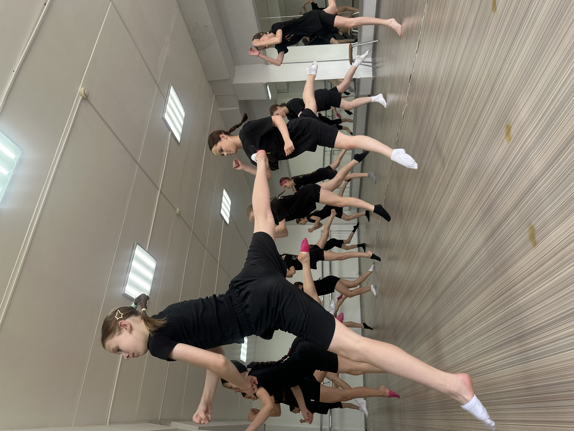 In a dance studio with mirrored walls and a slatted wooden wall, a group of young people in black dance attire are performing a synchronized routine. They are mostly wearing black shorts and black t-shirts or leotards, with various colored socks, including white, pink, and grey. The individuals are positioned at different heights, suggesting they are in mid-air or on elevated platforms, possibly as part of a complex choreography.
The lighting appears to be artificial, emanating from rectangular fluorescent panels on the ceiling. The overall scene evokes a sense of dedication and practice, with the dancers focused on their movements. The composition of the photo, with many legs and torsos visible, creates a dynamic and energetic impression. The mirrors reflect the dancers, doubling the visual impact of the performance and offering a sense of depth within the studio space. The image is a snapshot of a dance class or rehearsal, highlighting the athleticism and artistic expression of the participants. There is no visible text in the image. The time of day and weather are not discernible from the provided image.