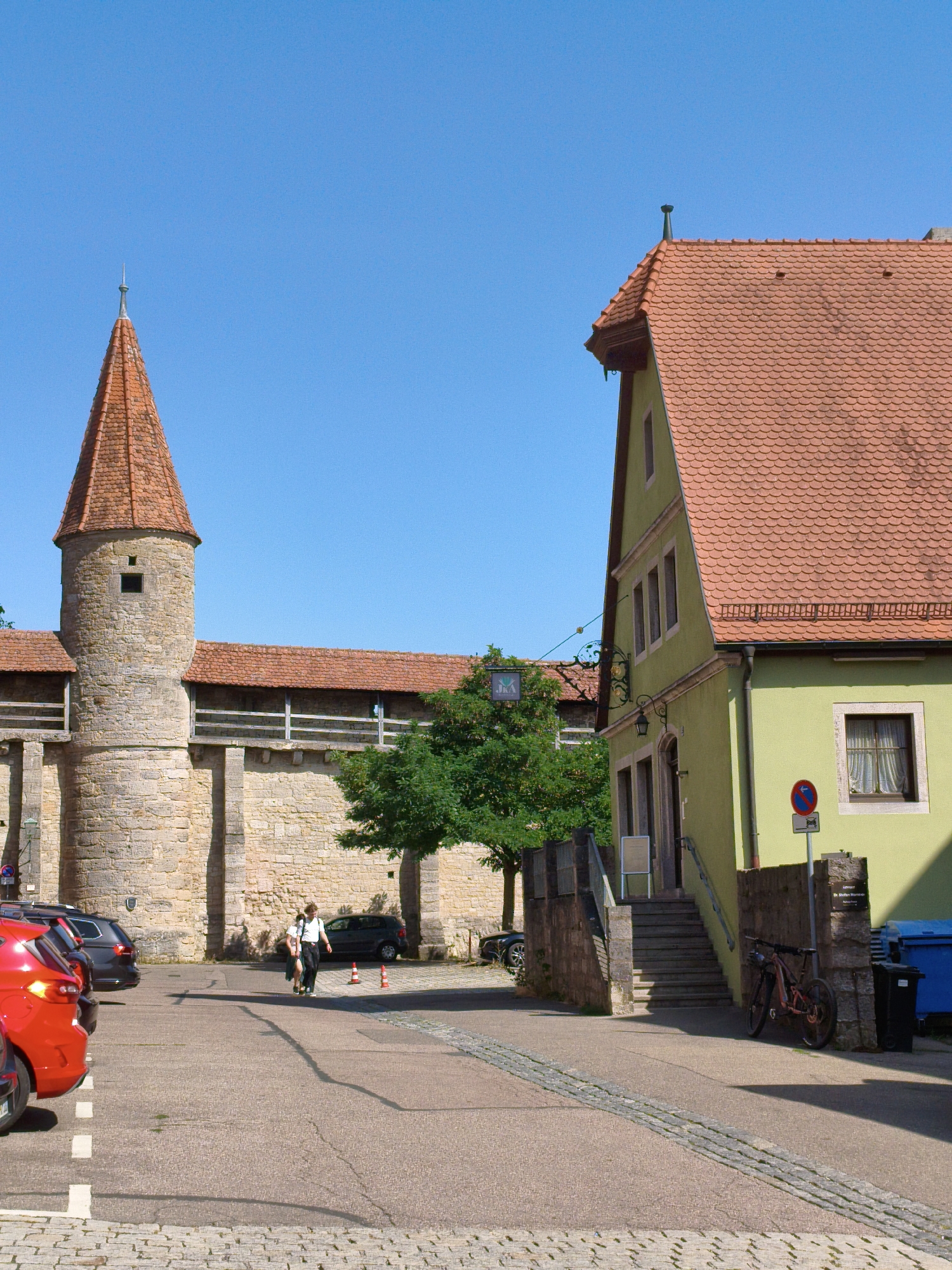 The image depicts a street scene in Rothenburg ob der Tauber, Germany. In the foreground, several cars are parked in a lot, including a bright red car. To the right, a bicycle leans against a stone wall. In the background, a medieval stone tower with a red tiled roof stands tall, part of the city's ancient fortifications. Adjacent to the tower is a light green building with a red tiled roof. Two people are walking in the middle ground, and a few trees are visible. The sky is clear and blue.