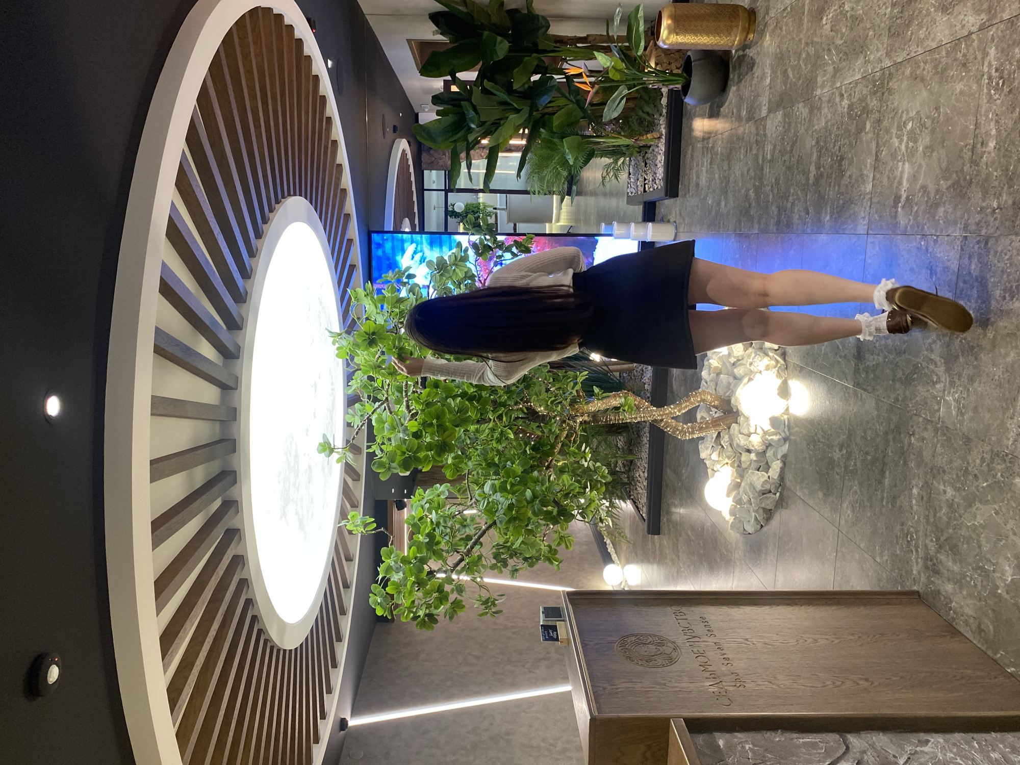 A woman, viewed from behind, walks away from the camera in a spa reception area. She has long, dark hair and wears a white long-sleeved shirt, a black skirt, white ankle socks with ruffles, and brown shoes. She reaches her right hand towards a decorative artificial tree with green leaves, some of which are illuminated by small lights. The tree is situated in a bed of white stones, with large, glowing white spheres placed around its base.
In the foreground to the left is a wooden reception desk with an embossed circular logo and text that reads "СЕДЬМОЕ ЧУВСТВО" and "Spa Seven Sense". The walls have a textured finish and are illuminated by vertical strip lights. Behind the woman, a large digital screen displays a colorful image, possibly of a natural landscape. To the right of the screen are several large potted plants, including a bird of paradise plant. The floor is tiled with a grey, marbled pattern. The ceiling features a circular design with concentric rings of wooden slats and a central luminous panel. The overall environment suggests a modern and tranquil spa setting. The location is identified as Votkinsk, Russia.