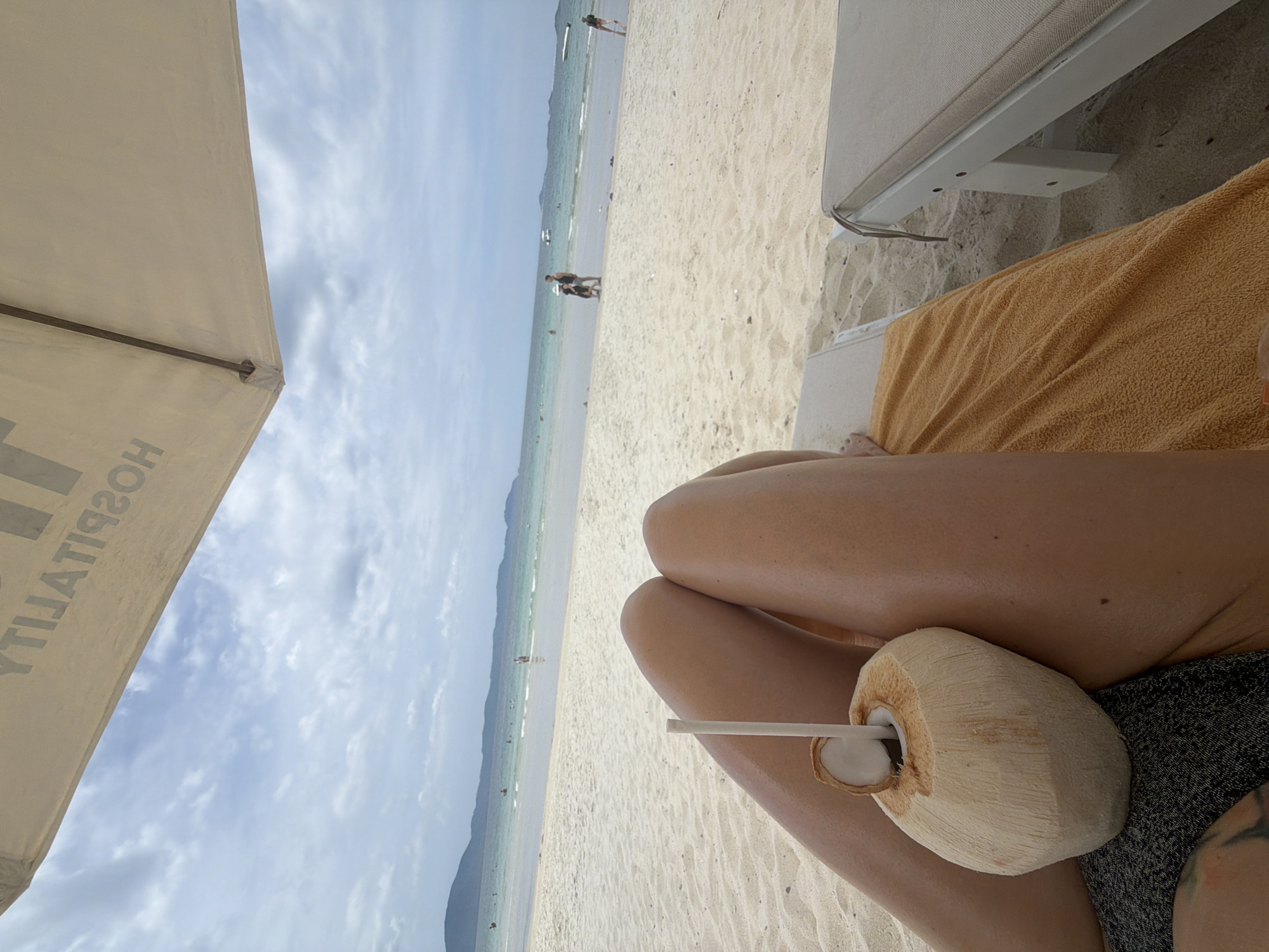 A first-person perspective shot captures a relaxing scene on a sandy beach, likely in Dong Ninh Hoa Ward, Vietnam. The foreground features a person's legs resting on a beach lounger, with a halved coconut with a straw positioned between their thighs. The coconut is partially covered by an orange towel. In the background, the ocean stretches to the horizon, with a line of mountains visible in the distance. Several people are scattered across the beach and in the shallow water, engaged in activities like swimming and walking. The sky is overcast with scattered clouds. A large, light-colored beach umbrella partially shades the scene, with faint lettering visible on its underside.
