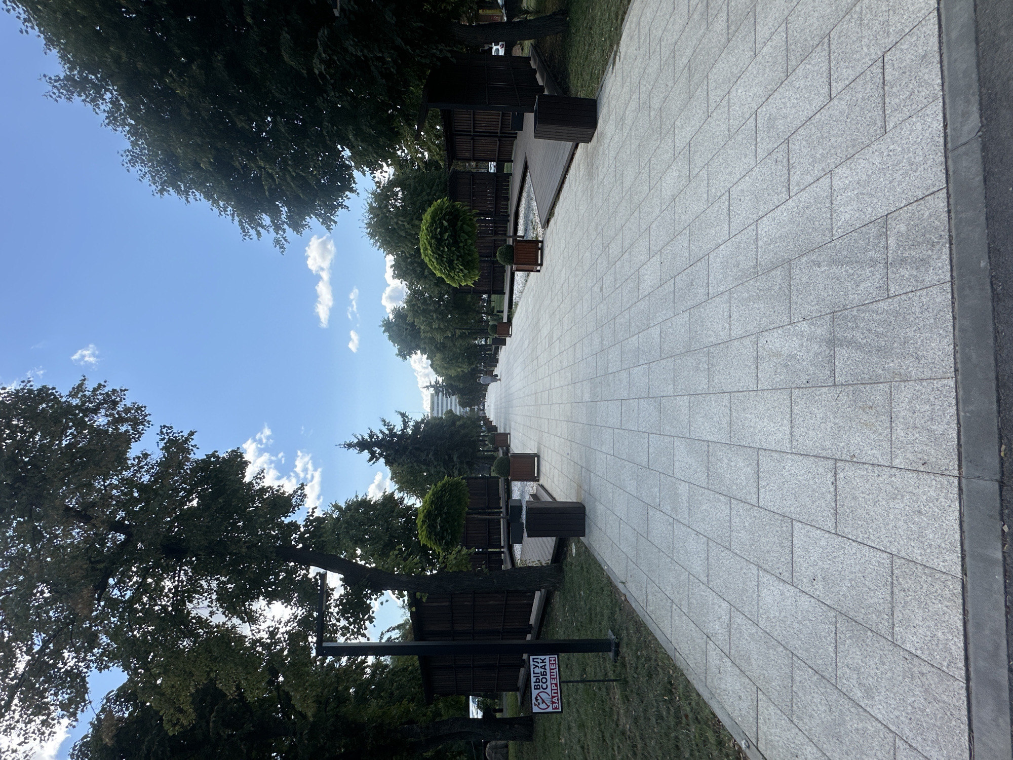 A wide, paved walkway, constructed from light gray granite tiles laid in a herringbone pattern, stretches into the distance under a partly cloudy blue sky. Tall, mature trees with dense green foliage line both sides of the walkway, casting shadows. On the left, a sign with a red circle and slash over a dog symbol, and the text "ВЫГУЛ СОБАК ЗАПРЕЩЕН" (Dog walking is prohibited) is posted on a dark pole. Adjacent to the walkway on the left are wooden gazebos with dark brown slatted walls and thatched roofs, and a grassy area. On the right, similar wooden structures with open sides and dark slatted roofs are present, along with planters containing small, manicured trees and bushes. In the background, a modern, white building with multiple floors is visible. A lone pedestrian, appearing as a distant figure, is walking away from the camera on the right side of the walkway. The overall scene depicts a well-maintained park or public square in Zhovte, Ukraine.