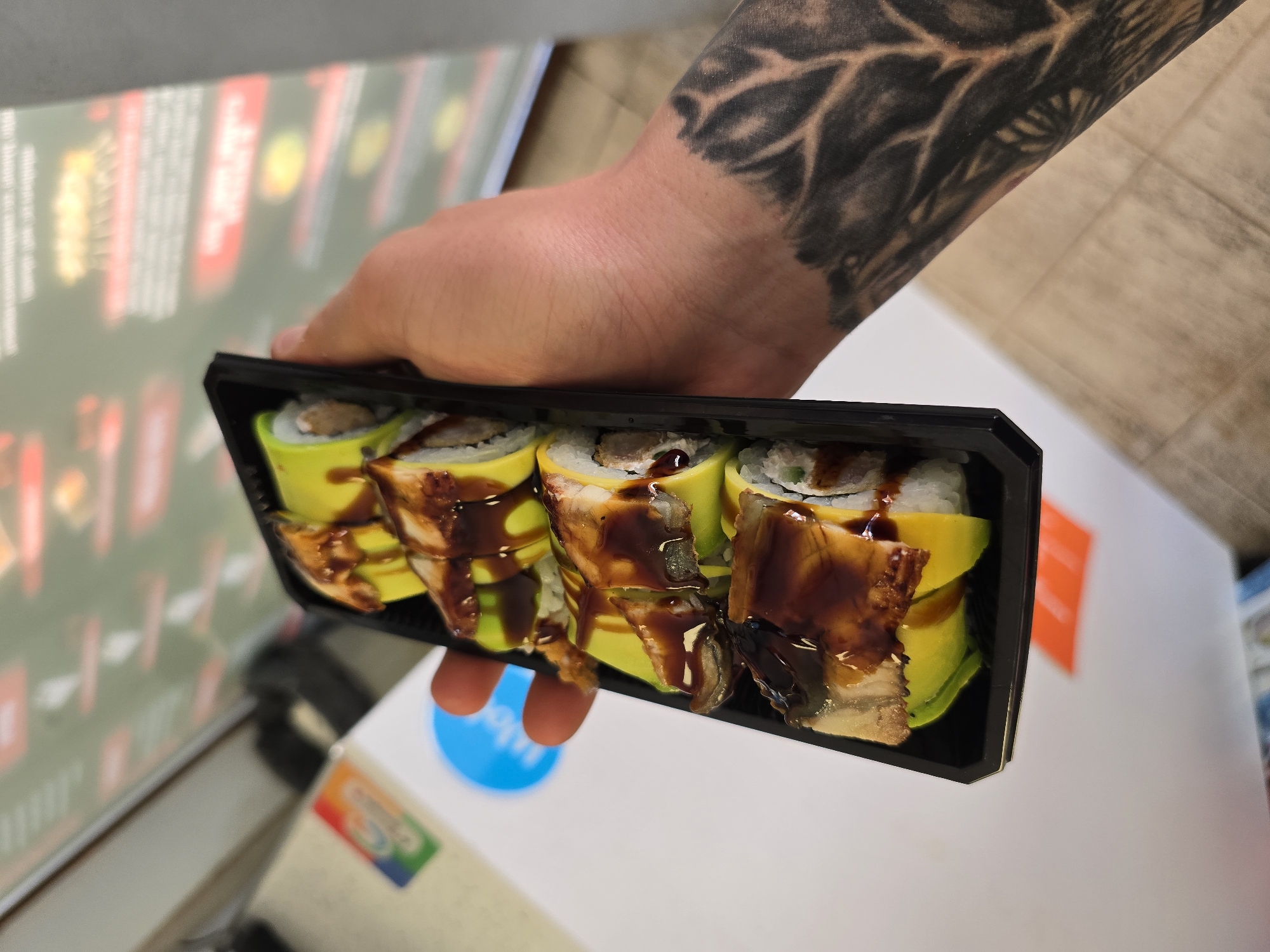 A person with a tattooed arm is holding a black container of sushi rolls. The sushi rolls appear to be topped with unagi (eel) and sauce, and wrapped with avocado. The background shows a blurred shelf with printed text in what appears to be a store. A blue and white sticker is visible on the surface beneath the sushi container. Based on the sticker and other potential indicators on the shelf in the background, the scene is likely in a shop or grocery store in Burgas, Bulgaria.