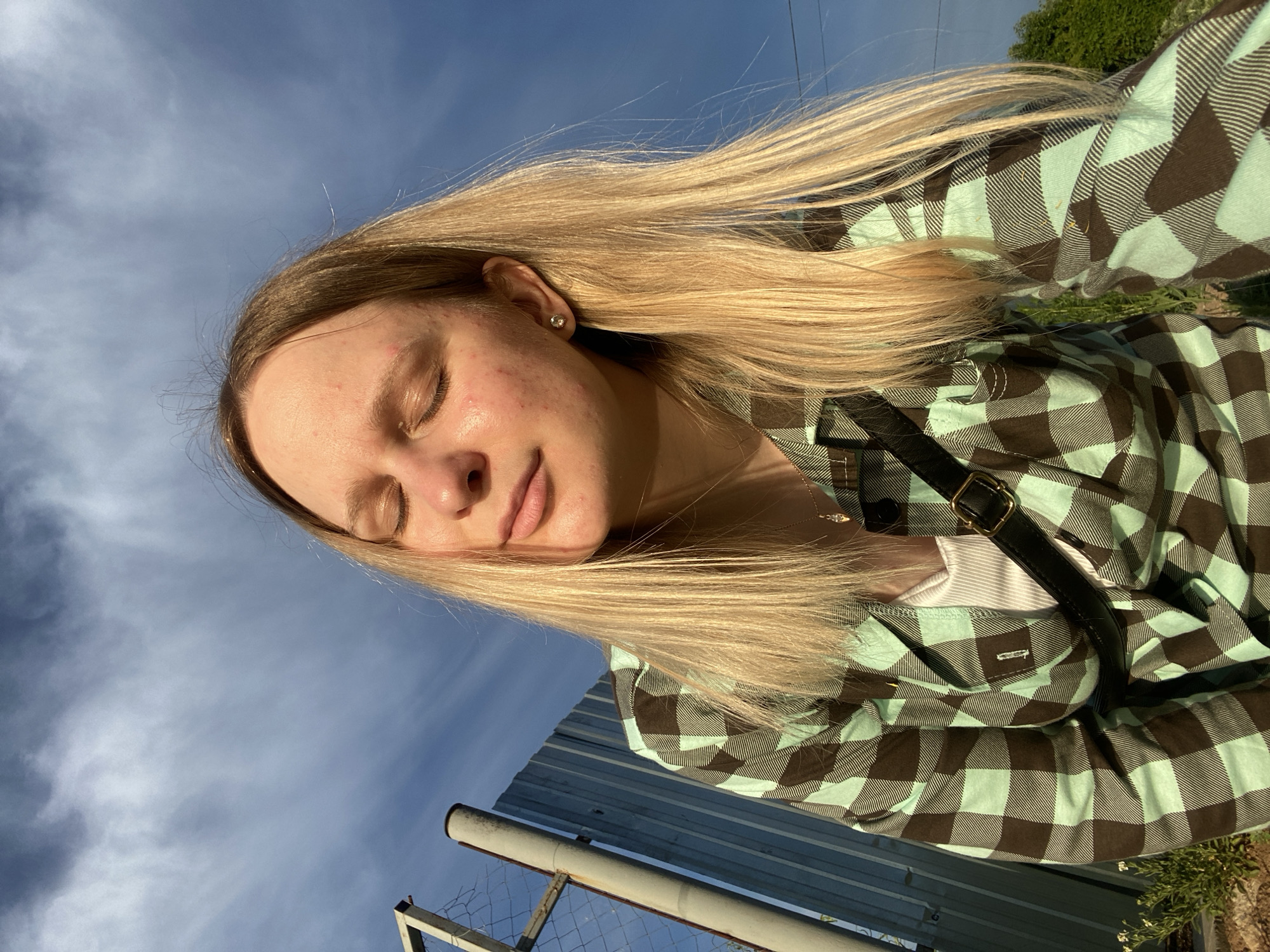 A young woman with long blonde hair is captured in a close-up, slightly tilted portrait, likely a selfie, outdoors. Her eyes are closed, and she wears a relaxed, peaceful expression with a faint smile, suggesting she is enjoying the sunlight. Visible blemishes are present on her forehead and right cheek. She is dressed in a mint green and brown plaid long-sleeved shirt, worn over a white inner top. A black strap, presumably from a bag, crosses her chest, featuring a gold buckle. A small gold stud earring is visible in her right ear.
The background consists of a clear blue sky with wispy white clouds, indicating a sunny day. The warm, golden light illuminates her face, suggesting the photo was taken during late afternoon or early evening. Below her, a corrugated metal fence or structure and a metal post are visible, hinting at an urban or industrial-edge outdoor setting. Some green foliage is present in the upper right background, and faint power lines can be seen against the sky. There is no visible text in the image. The scene conveys a sense of calm and natural light.