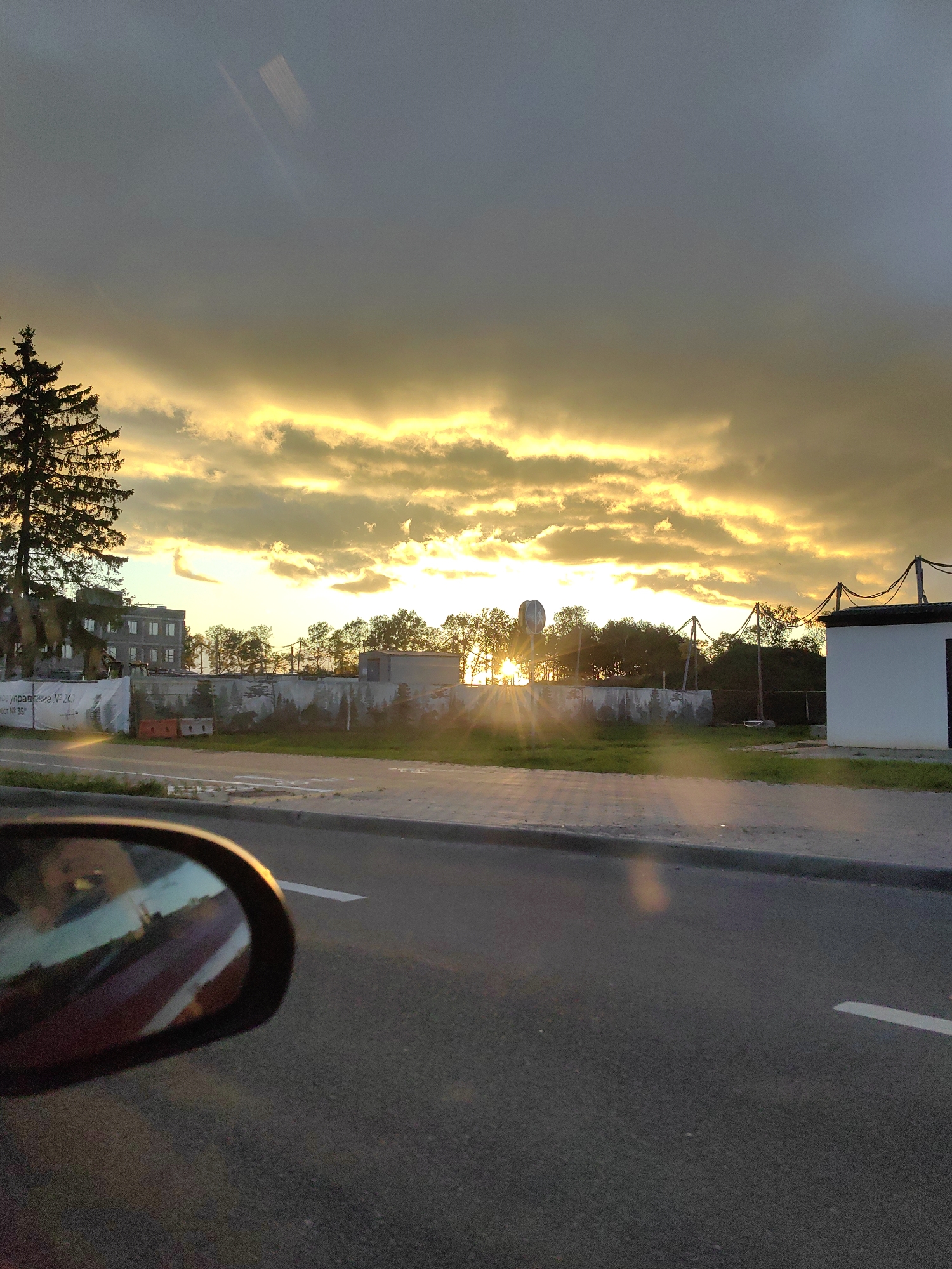 A low-angle, medium shot captures a view from a vehicle traveling down a paved road in Fanipal, Belarus. The scene is dominated by a dramatic sunset with golden light streaming through dark, dramatic clouds, casting a warm glow across the sky. In the foreground, the side mirror of the vehicle reflects a red interior. The road features a grey curb and a white lane marking. Beyond the road, a grassy median separates it from a fenced-off area. A tall evergreen tree stands to the left, silhouetted against the bright sky. Behind the fence, various structures are visible, including a multi-story building, trees, and a small white building with a black roof on the right. The overall atmosphere is serene, with the vibrant sunset providing a striking backdrop to the urban landscape.